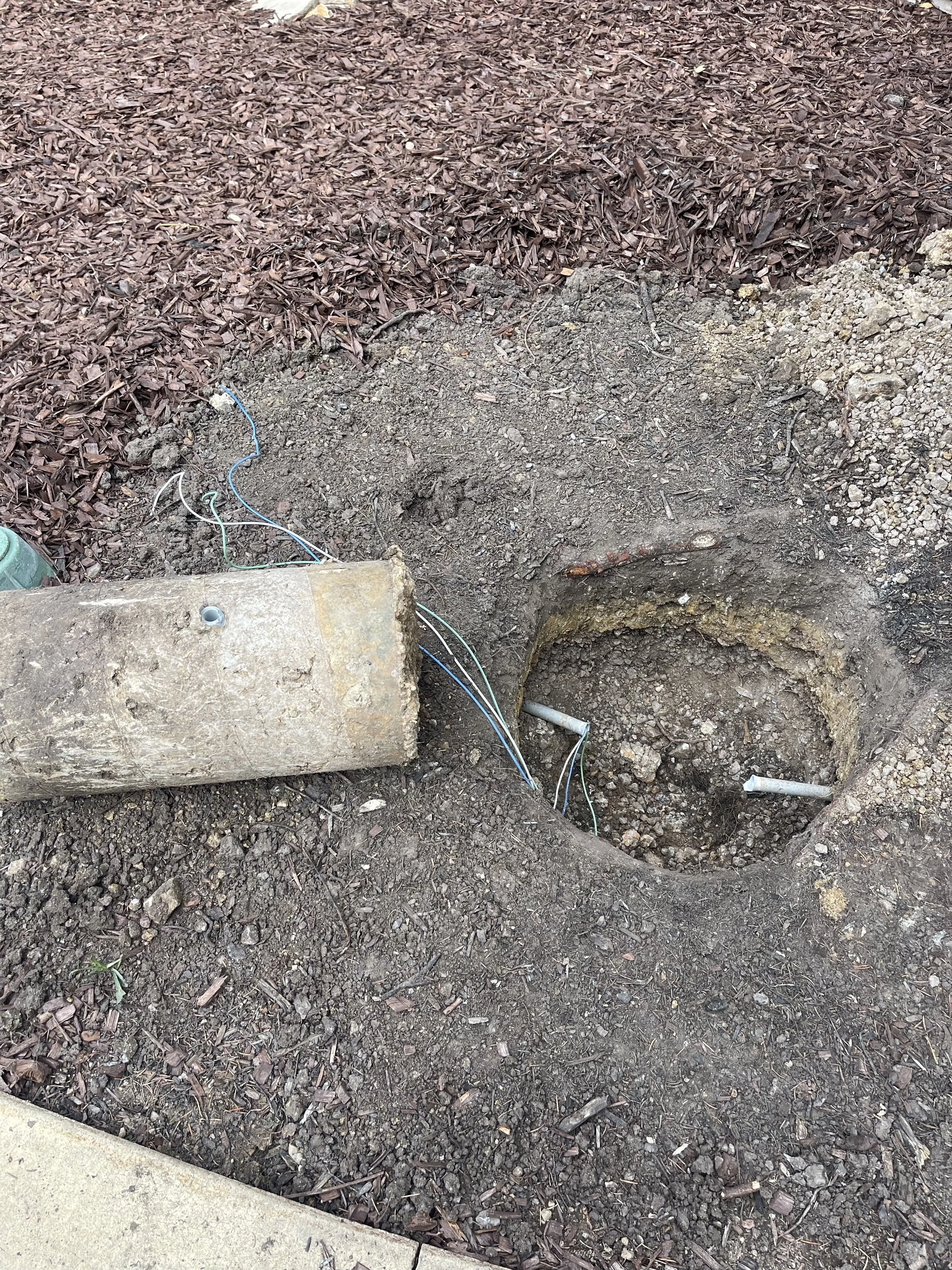 An open hole in the ground with exposed electrical wires and pipes. There is a concrete block nearby and mulch covering the surrounding area.