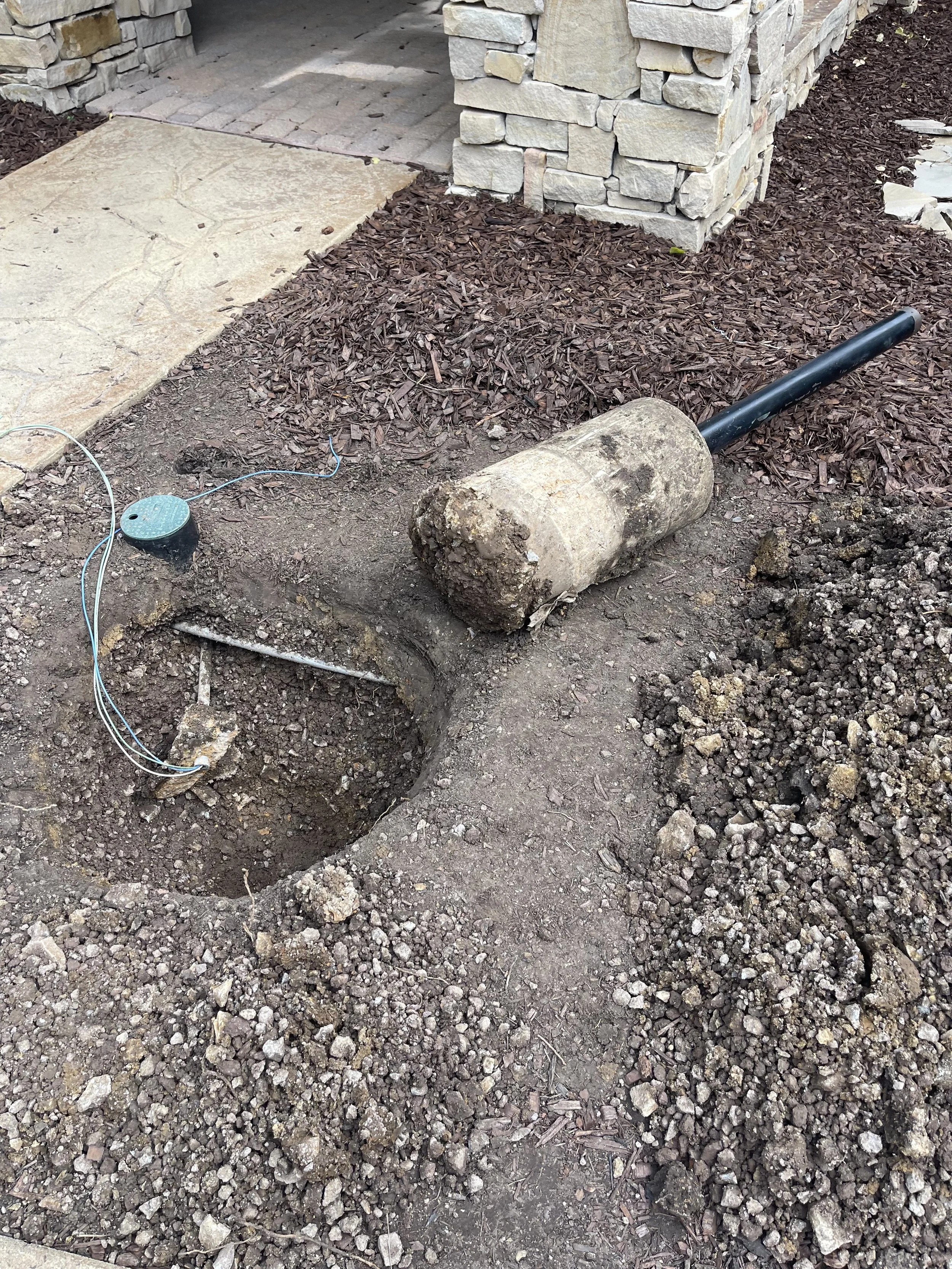 A hole in the ground with a large concrete mallet and some wires nearby, indicating a construction or landscaping project.