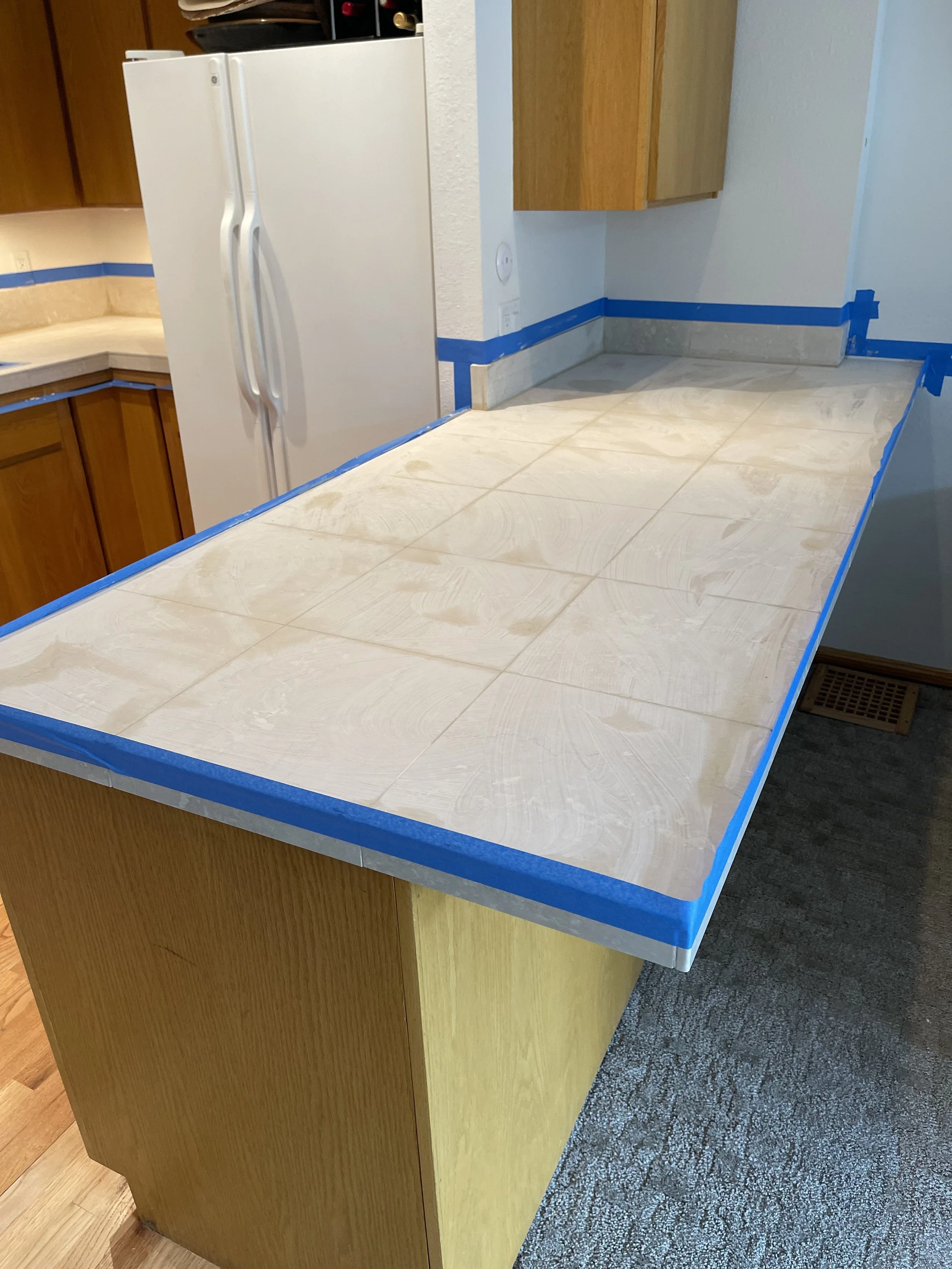 Kitchen countertop with tiles in progress, blue painter's tape along the edges, wooden cabinets, white refrigerator, and part of a carpeted floor.
