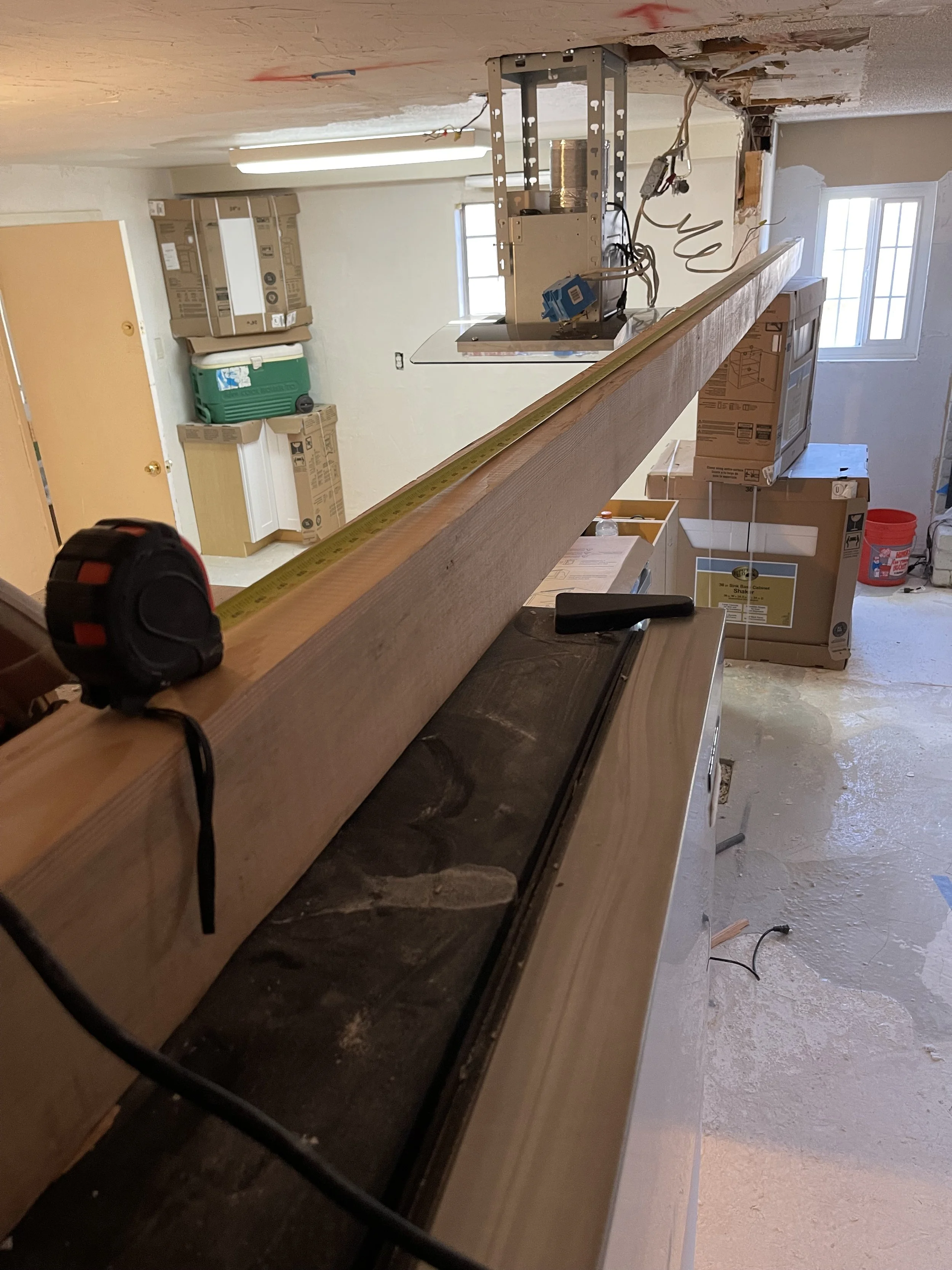 Construction site with a long wooden beam on a work surface, a measuring tape, and tools. The room is unfinished with boxes and construction materials, including a window and a ceiling with exposed framing.