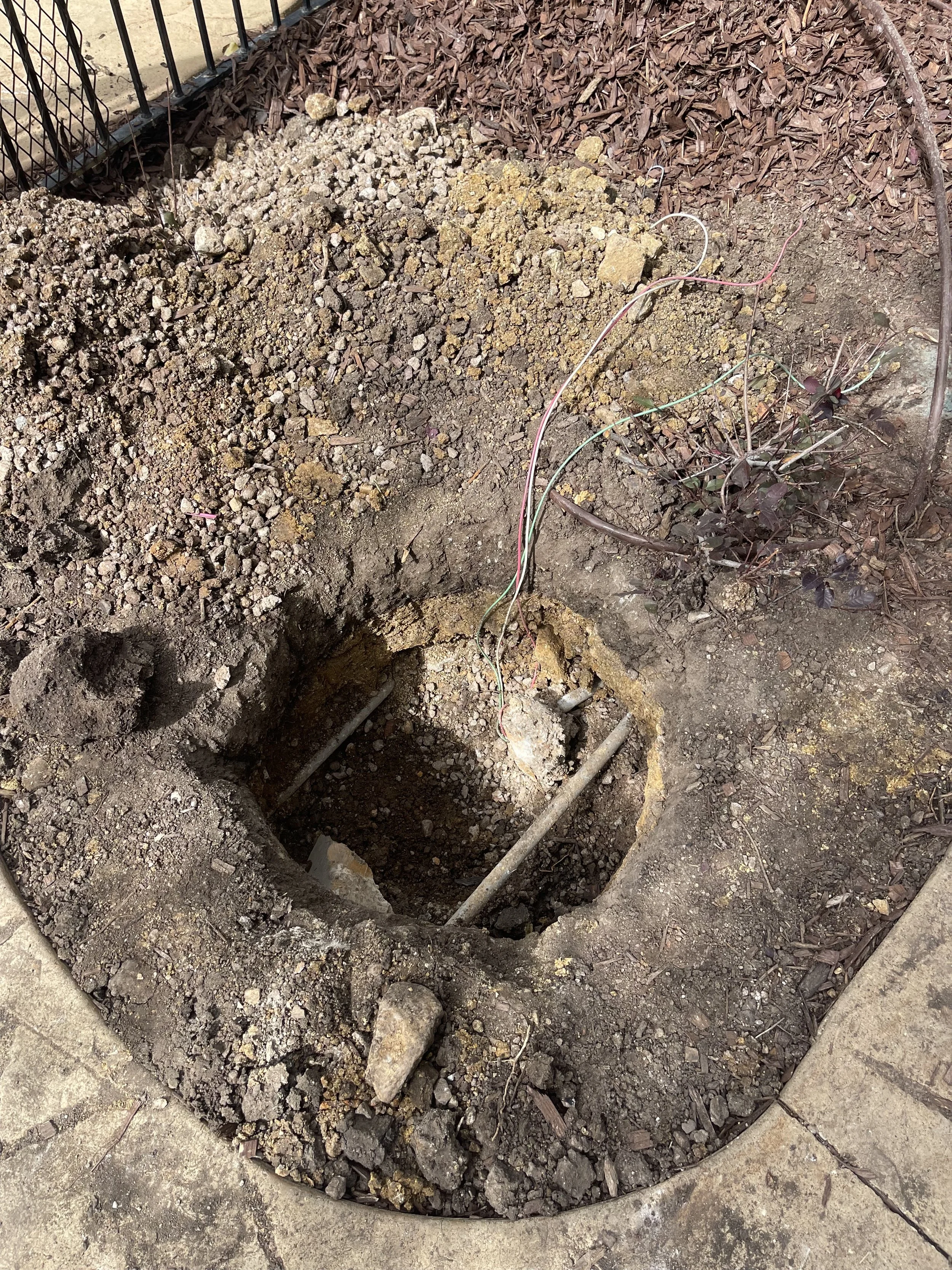 A hole dug in the ground with some exposed soil, a pipe, and wiring visible inside. Surrounding the hole are dirt, mulch, and a small shrub. A black metal fence is in the background.