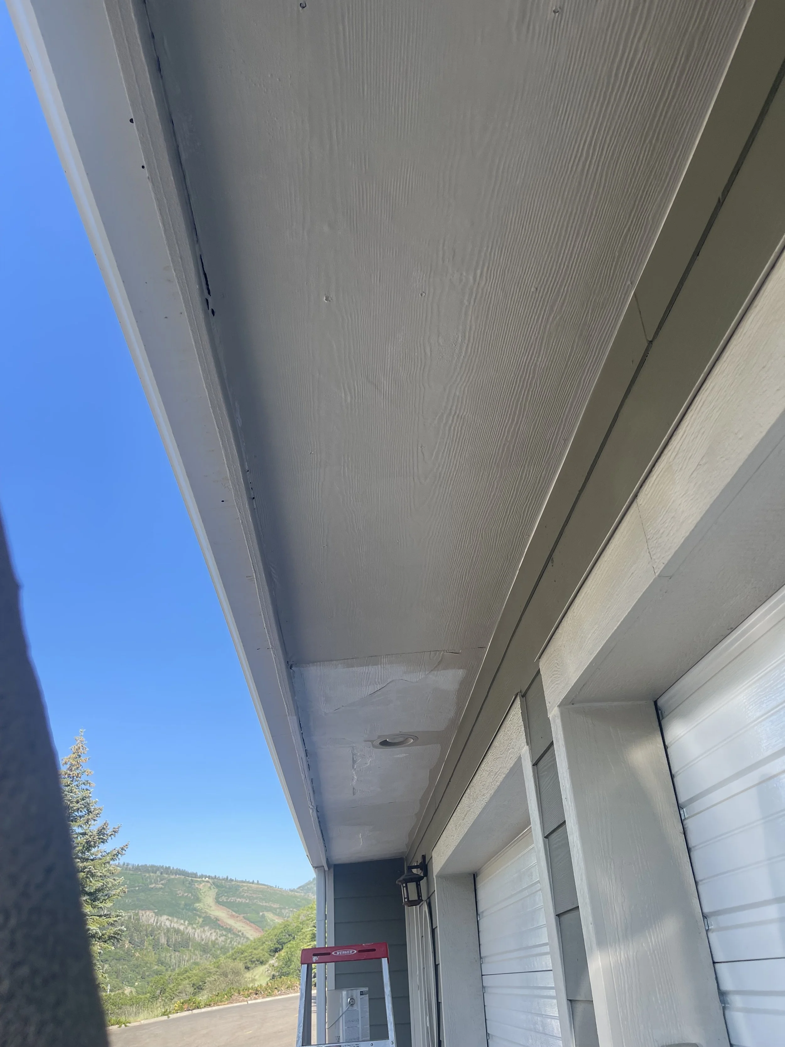 View of a house's exterior ceiling and part of a garage door with a scenic mountain landscape in the background. There is a step ladder and a wall lamp near the house.