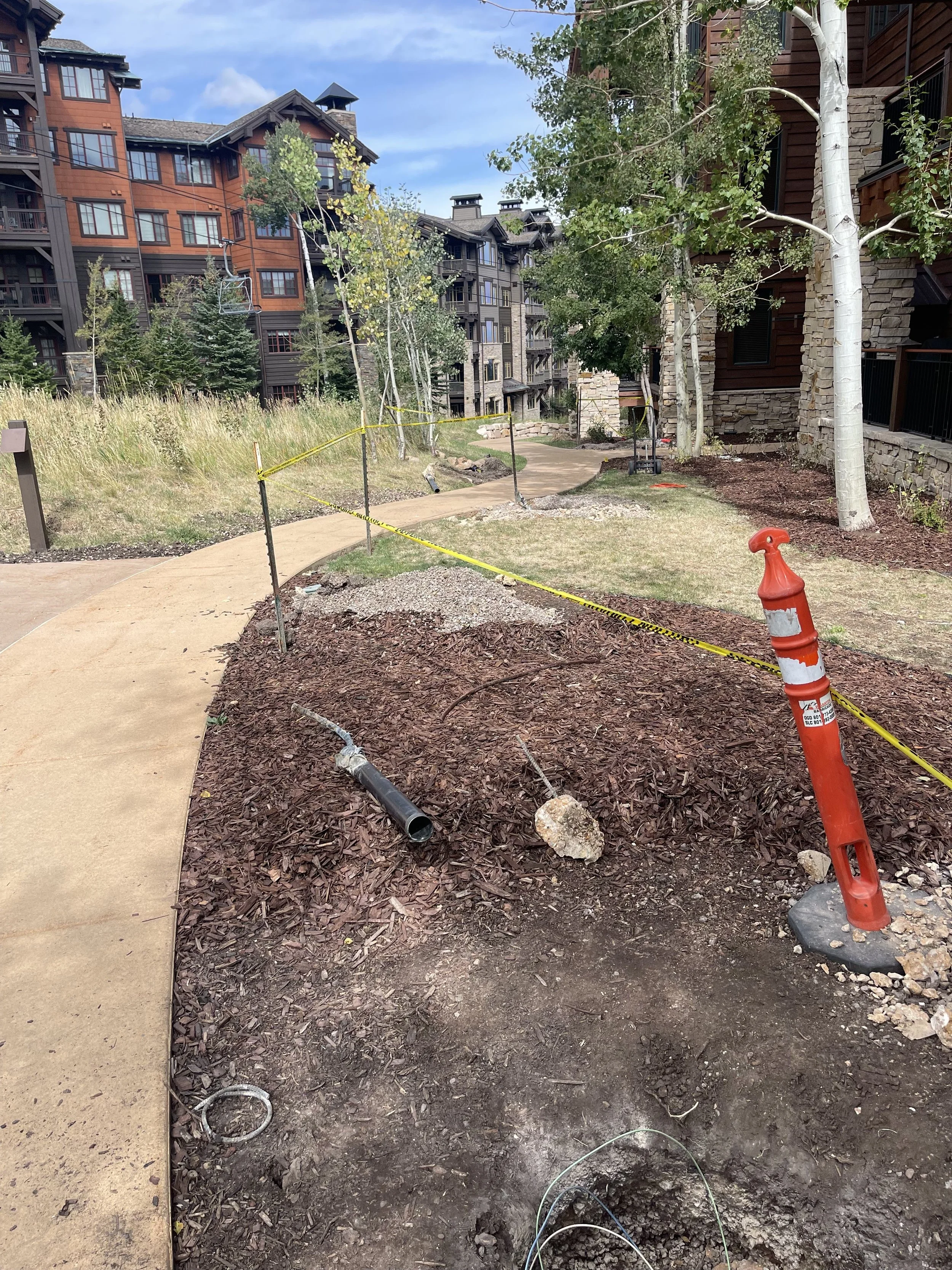 Sidewalk and landscaping area under construction in a residential neighborhood with multi-story buildings, trees, and yellow caution tape.