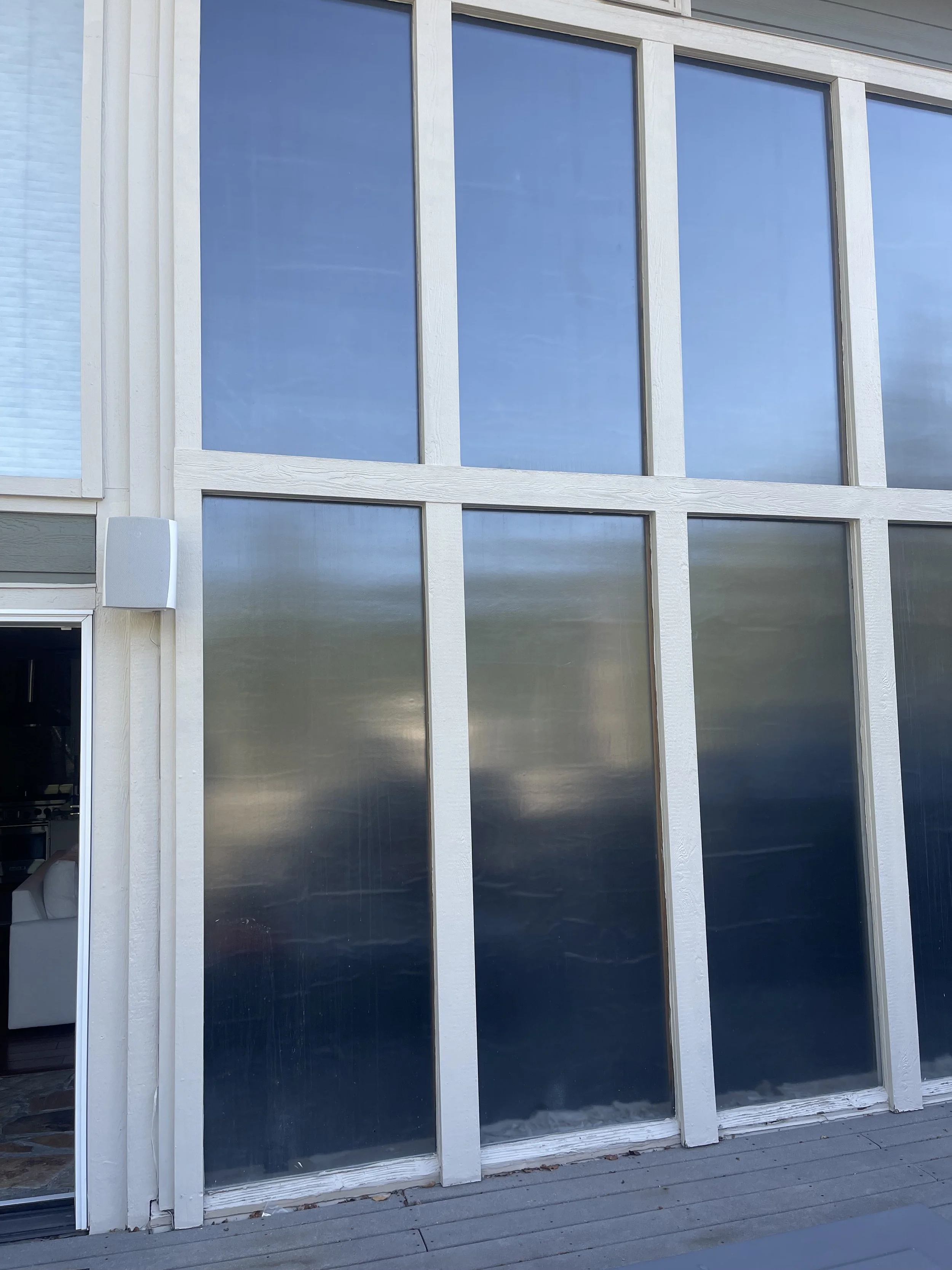 Large glass windows on a modern house exterior, with white framing and a sliding glass door on the left, reflecting the sky.