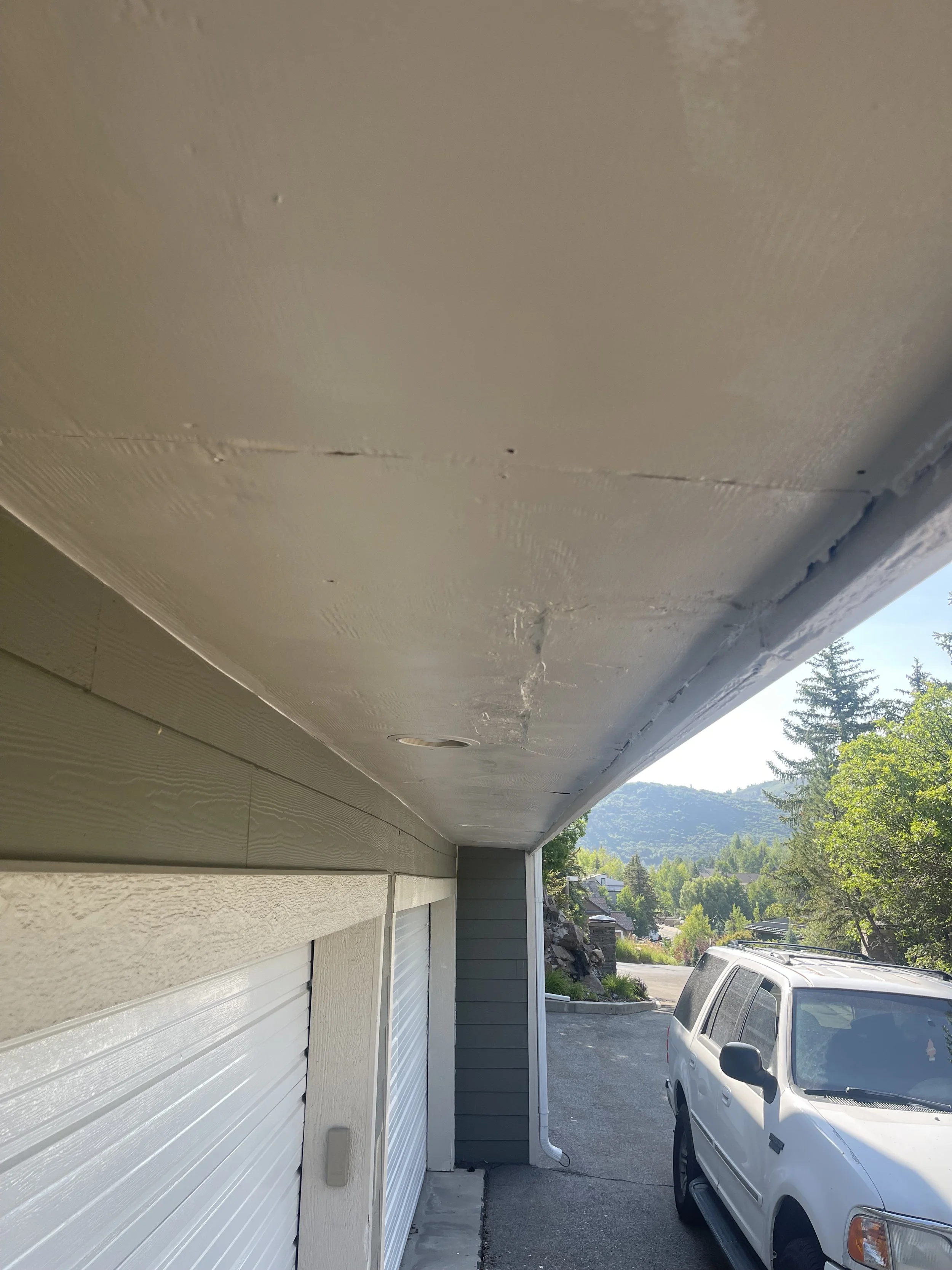 View of a garage roof with damage and cracks, adjacent to a parked white vehicle, with trees and mountains in the background on a sunny day.