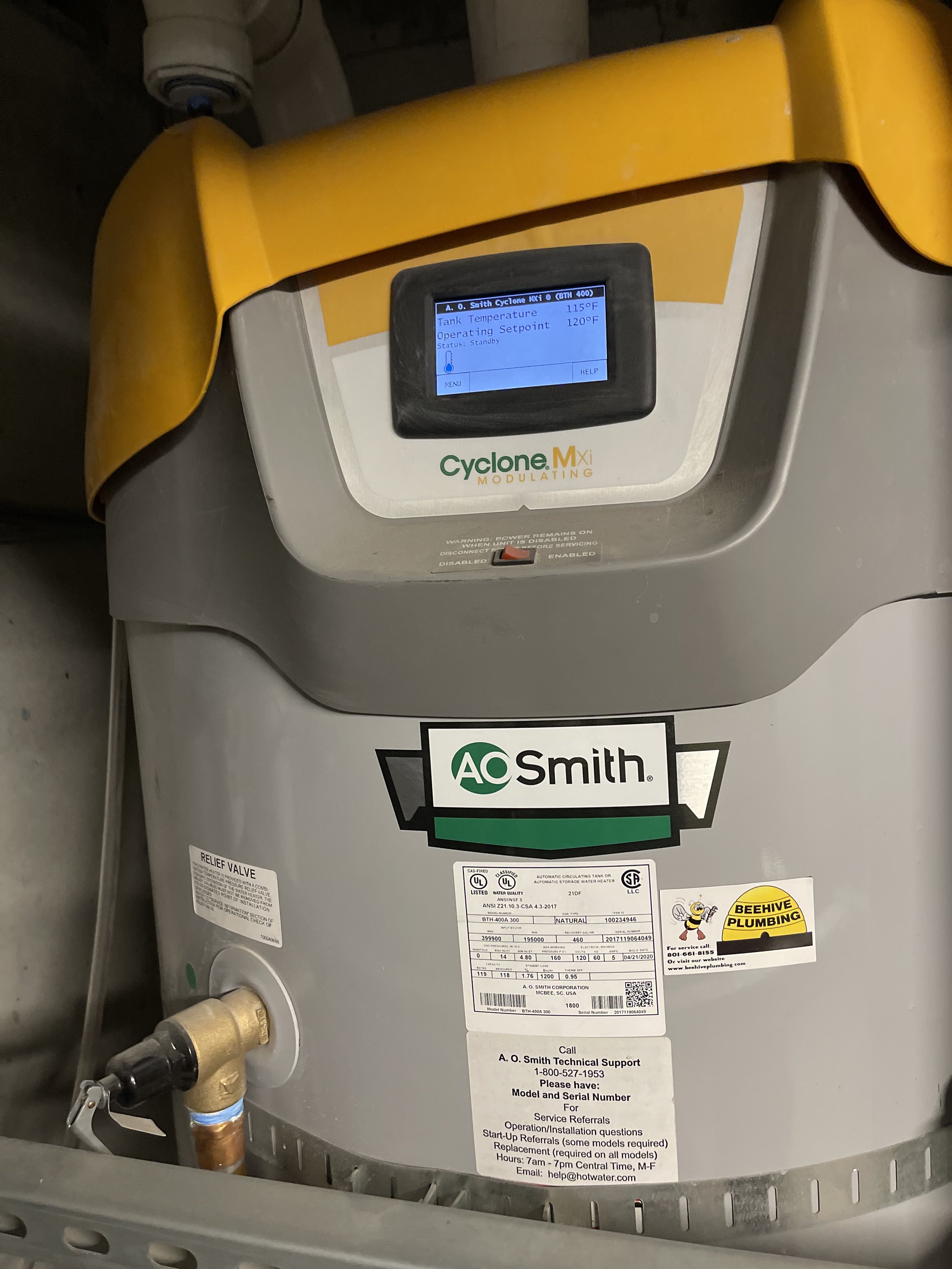 A large water heater with a digital control panel displaying temperature information, labeled A. O. Smith Cyclone MxI, and various labels including AC Smith and Beehive Plumbing.
