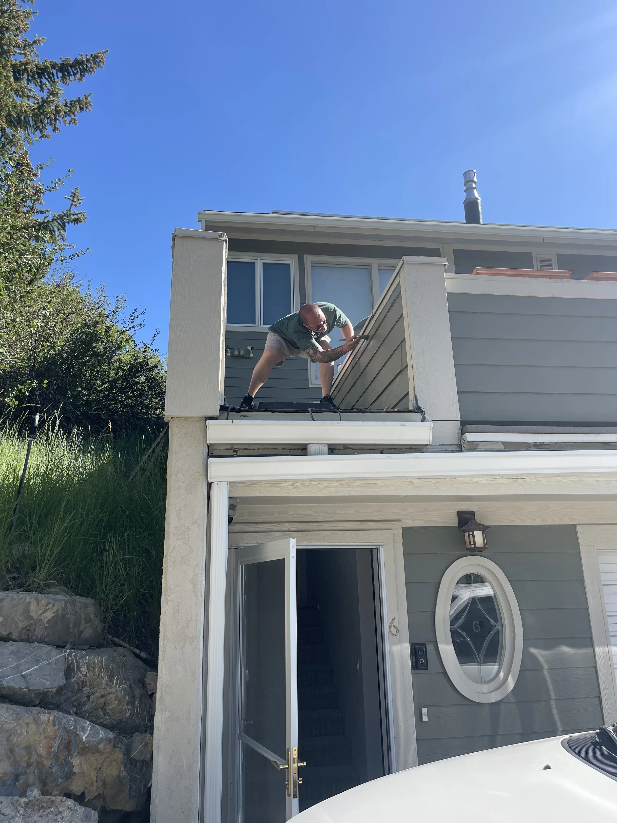 Man working on a house's exterior, leaning over the railing of the upper level, with an open door and a staircase visible below. The sky is clear and blue.