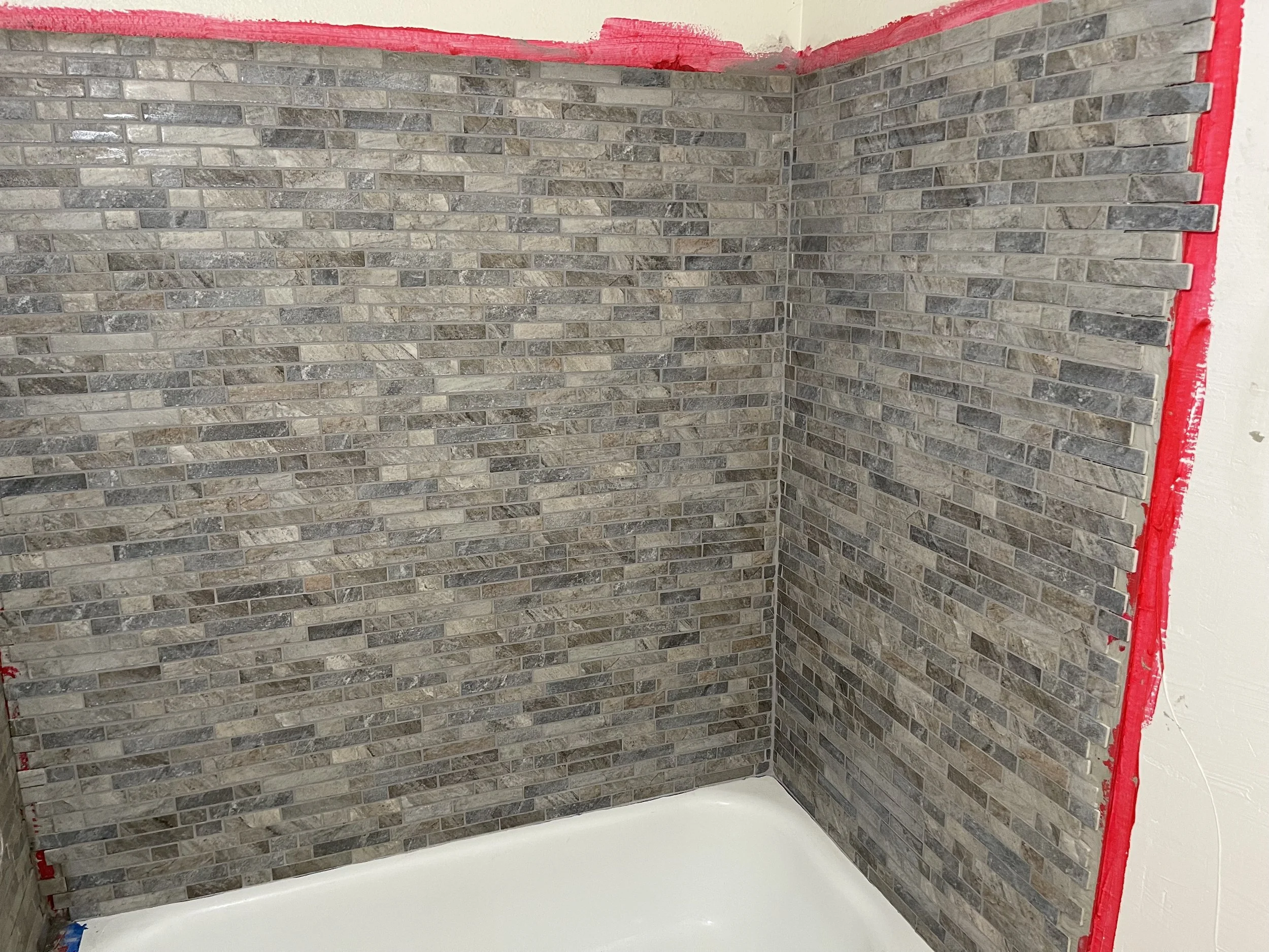 Corner of a bathroom shower area with multi-color gray brick tiles on the walls. Red painter's tape is along the edges, and there is a white shower pan at the bottom.