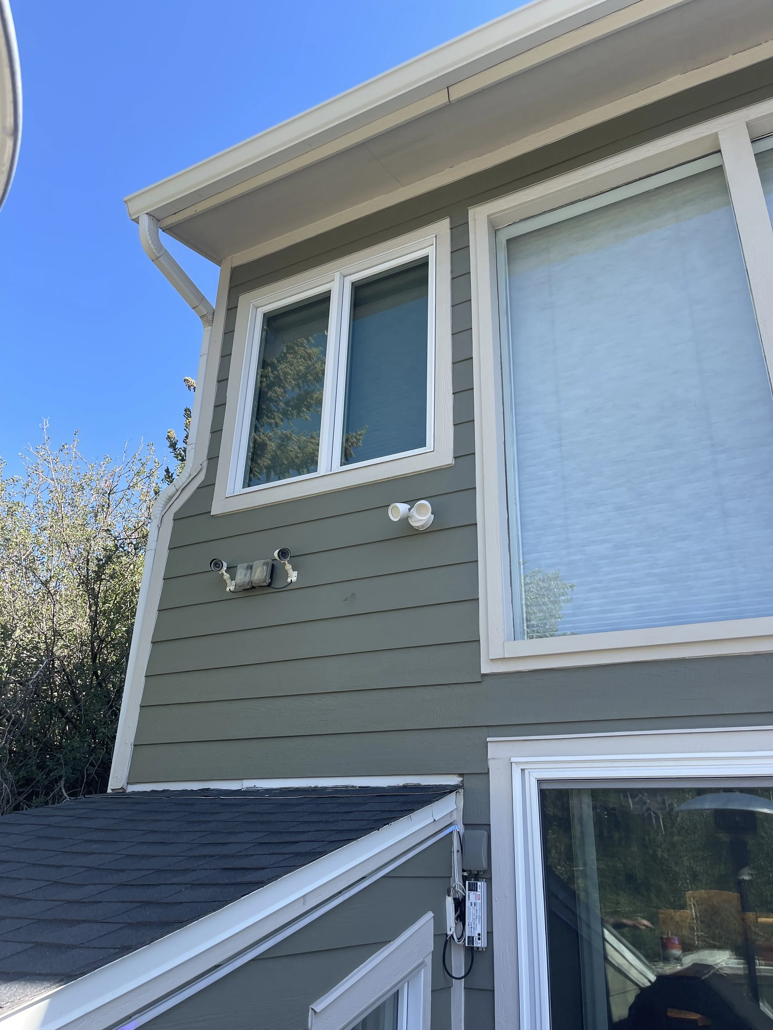 Close-up of the side of a house with grey siding, white trim, two large windows, surveillance cameras, and external wiring under a clear blue sky.