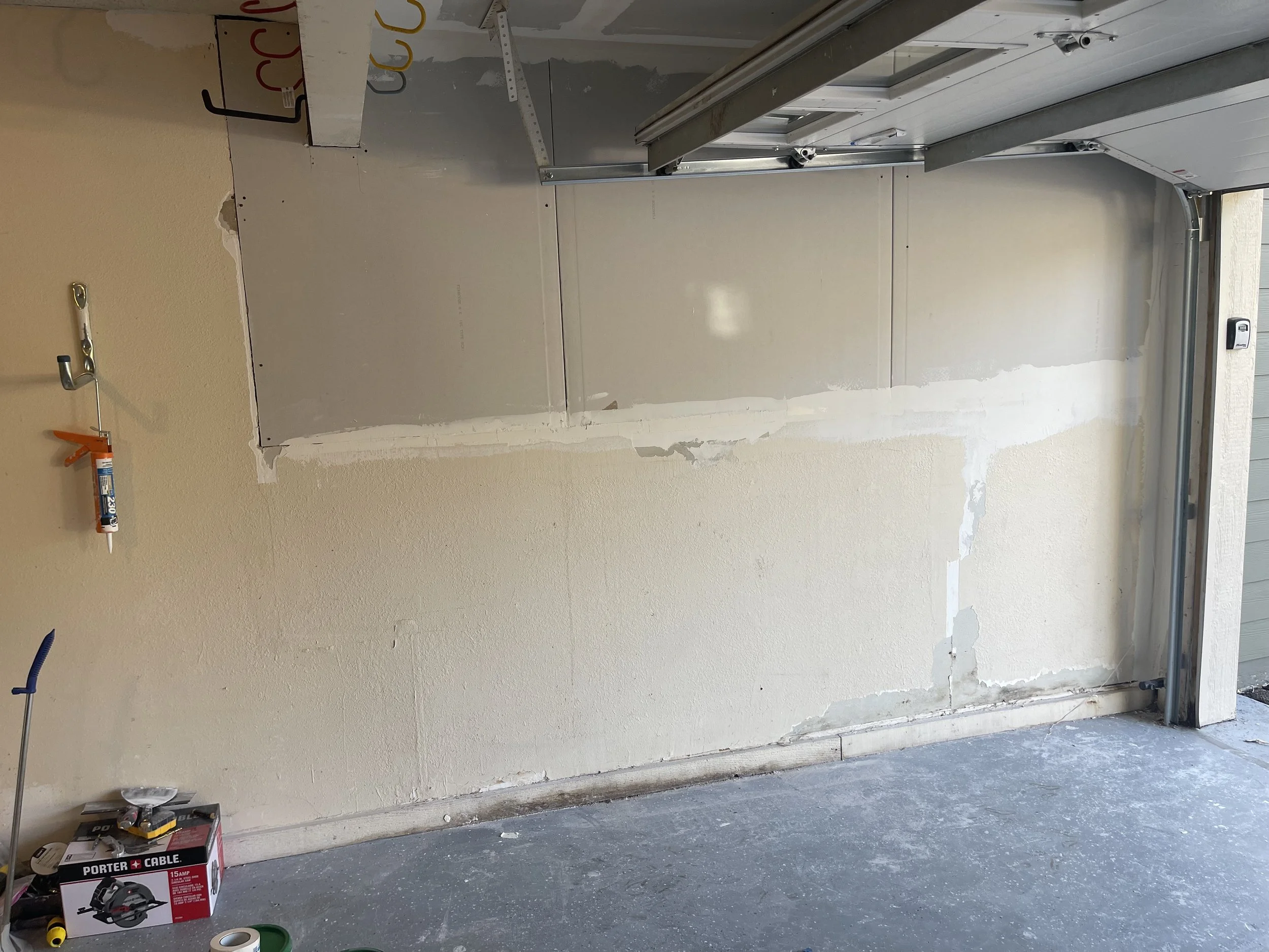 A garage wall with drywall prep for painting or finishing, showing partial drywall installation and joint compound application, with various tools and supplies on the floor.