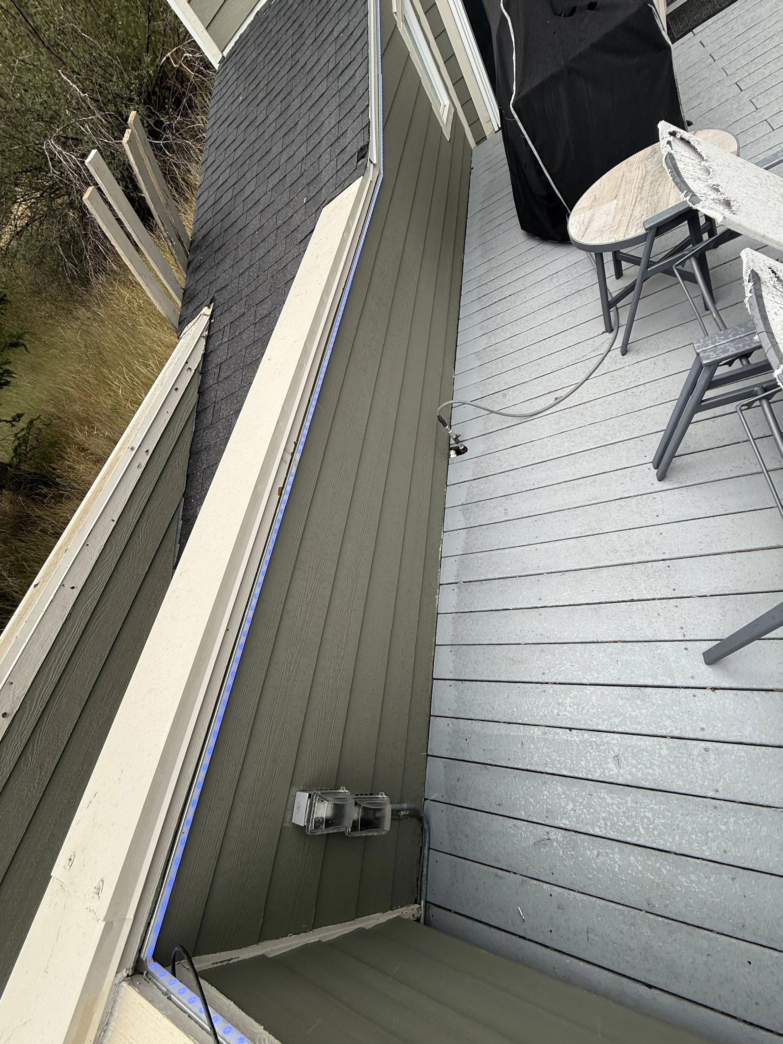 A rooftop deck with a grey wooden floor, outdoor chairs, and a covered grill. The deck is next to a house with green siding, black roof shingles, and a window. There are electrical outlets and wiring along the house exterior, with trees and a grassy 