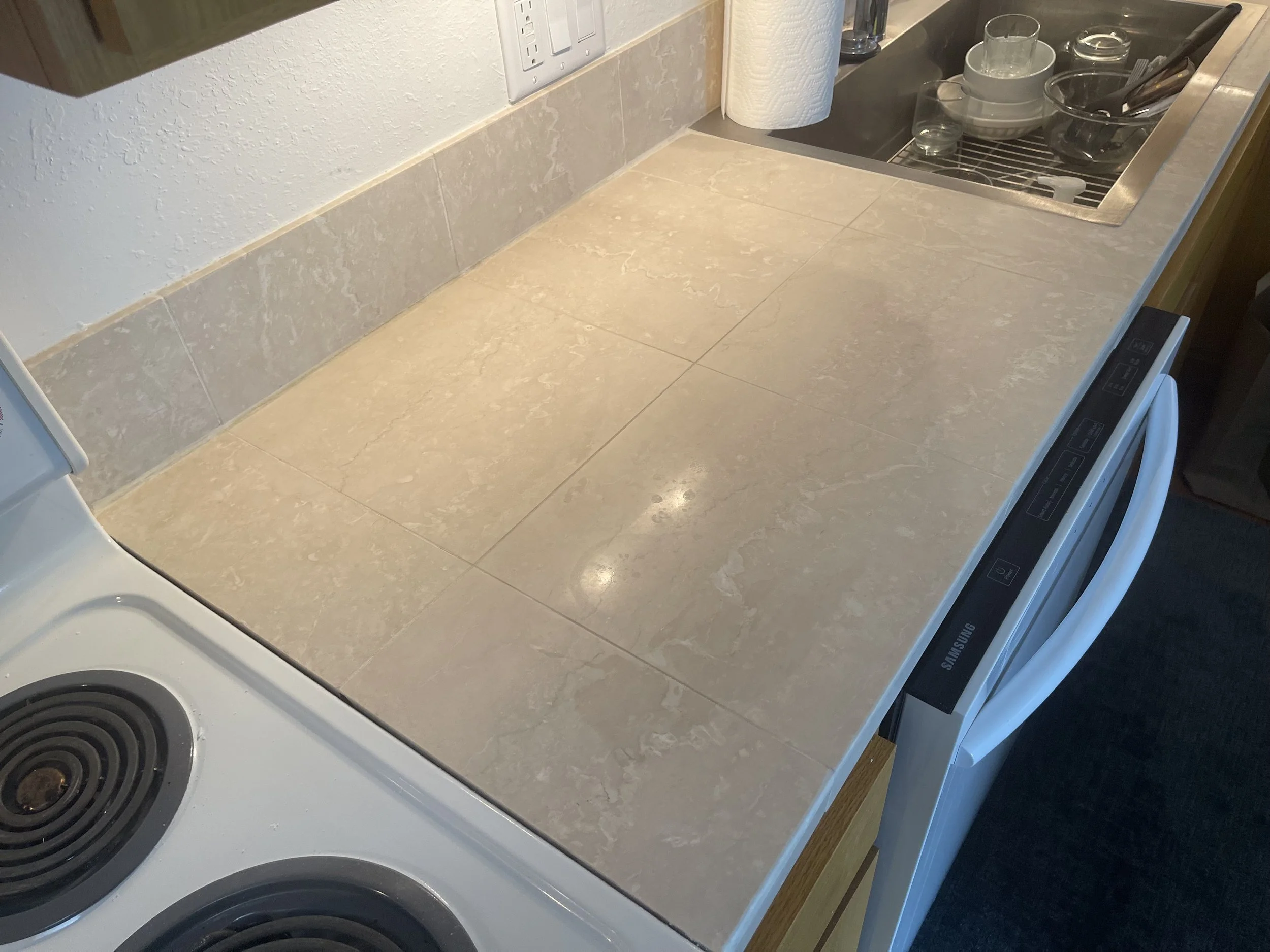 Kitchen counter with beige marble tiles, near a stove and a sink with dishes, glasses, and utensils.