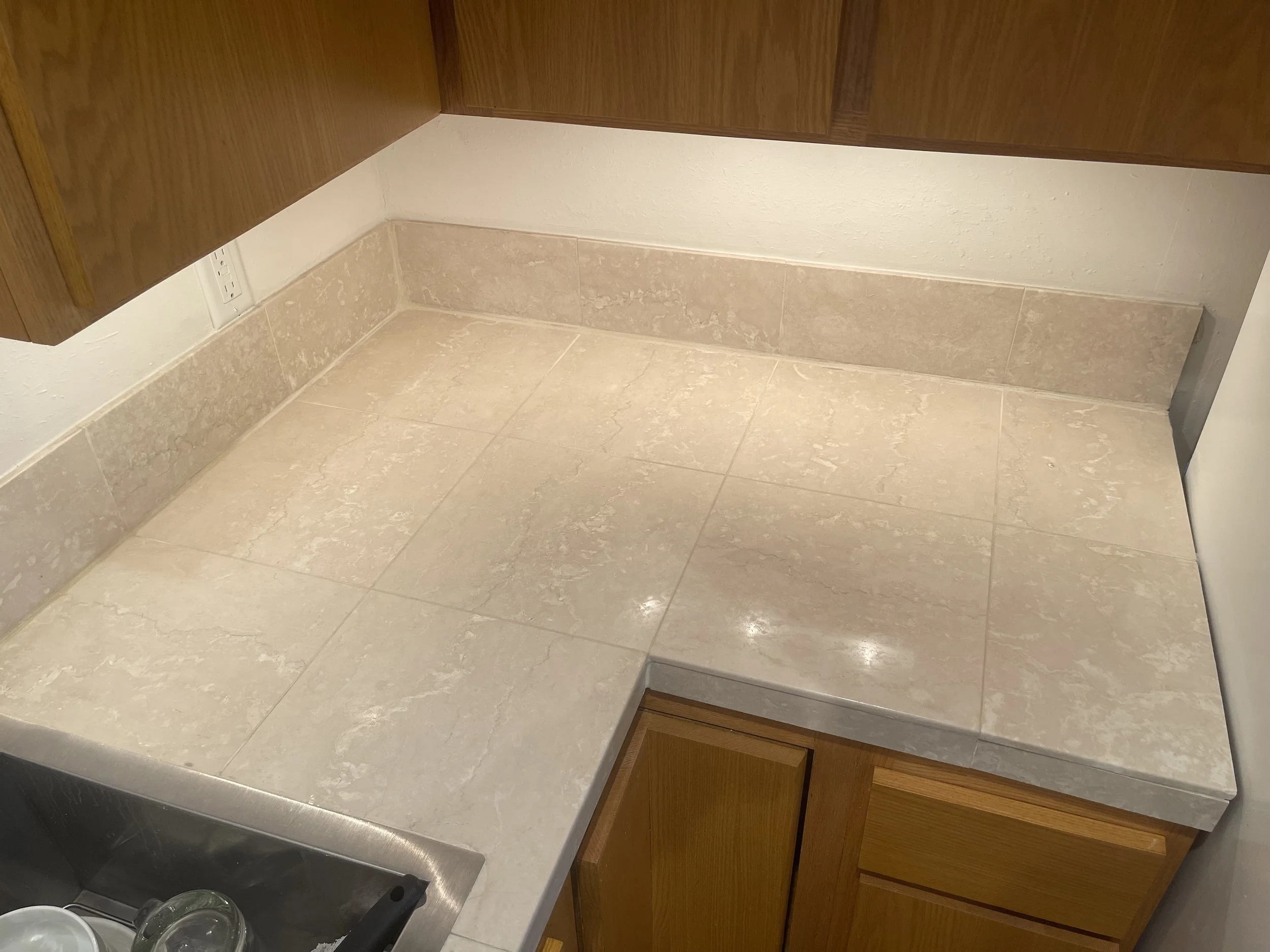 Kitchen countertop with beige marble tiles, a wooden cabinet above, and a sink with glassware on the left.