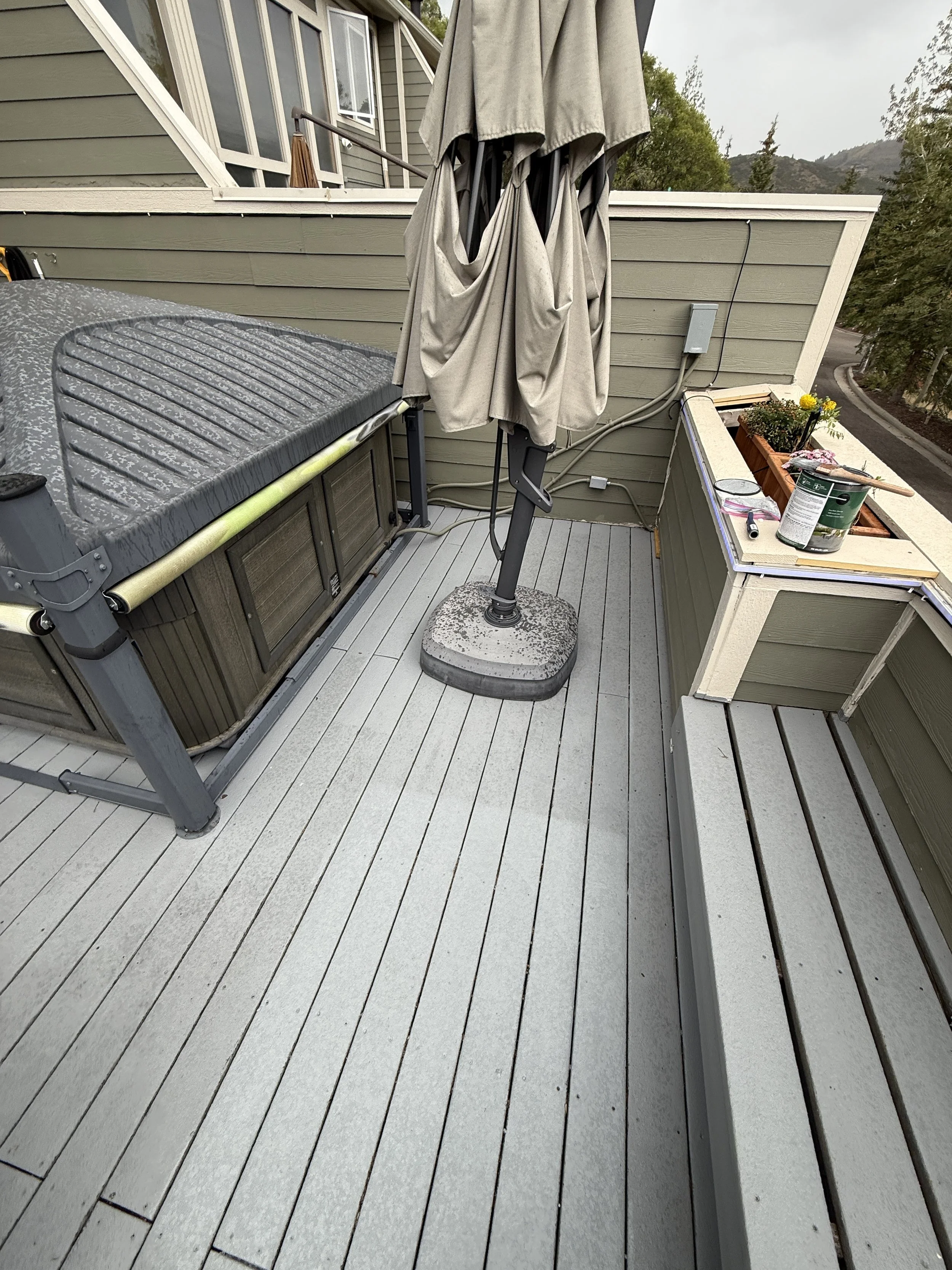 A rooftop deck with a hot tub, a closed patio umbrella, a planter box with flowers, and some gardening supplies. The deck has gray wooden flooring and green siding on the walls.