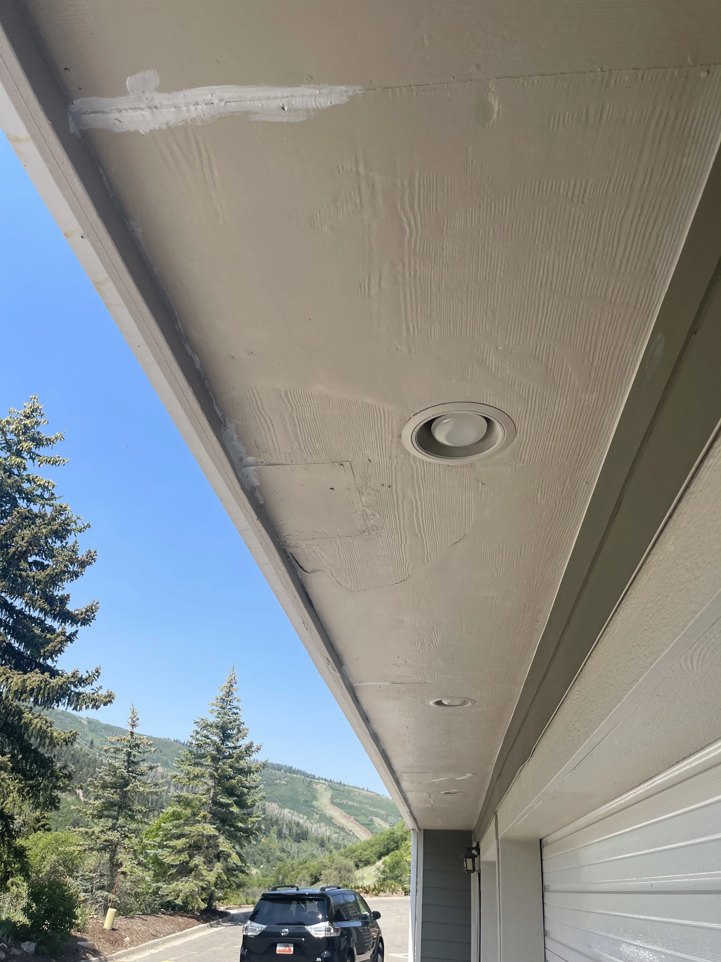 Close-up of a beige exterior house soffit with a recessed light and some visible patch repairs, with a parking lot, trees, and mountains in the background.