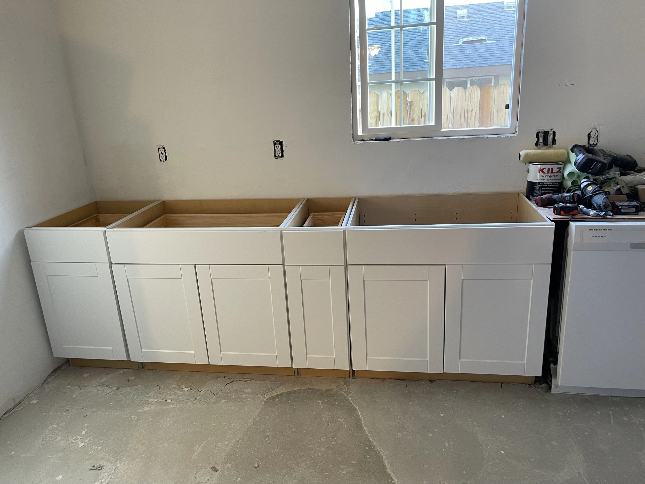 Kitchen cabinets under construction, with white cabinet doors and wooden interiors, placed under a window in a room with unfinished flooring and visible electrical outlets.