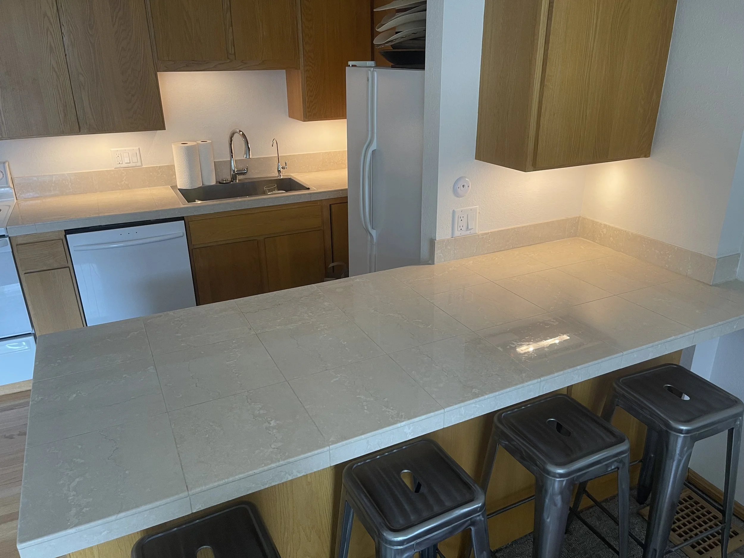 Kitchen with wooden cabinets, a white refrigerator, a white dishwasher, a beige counter with a sink, and three metal bar stools at a counter