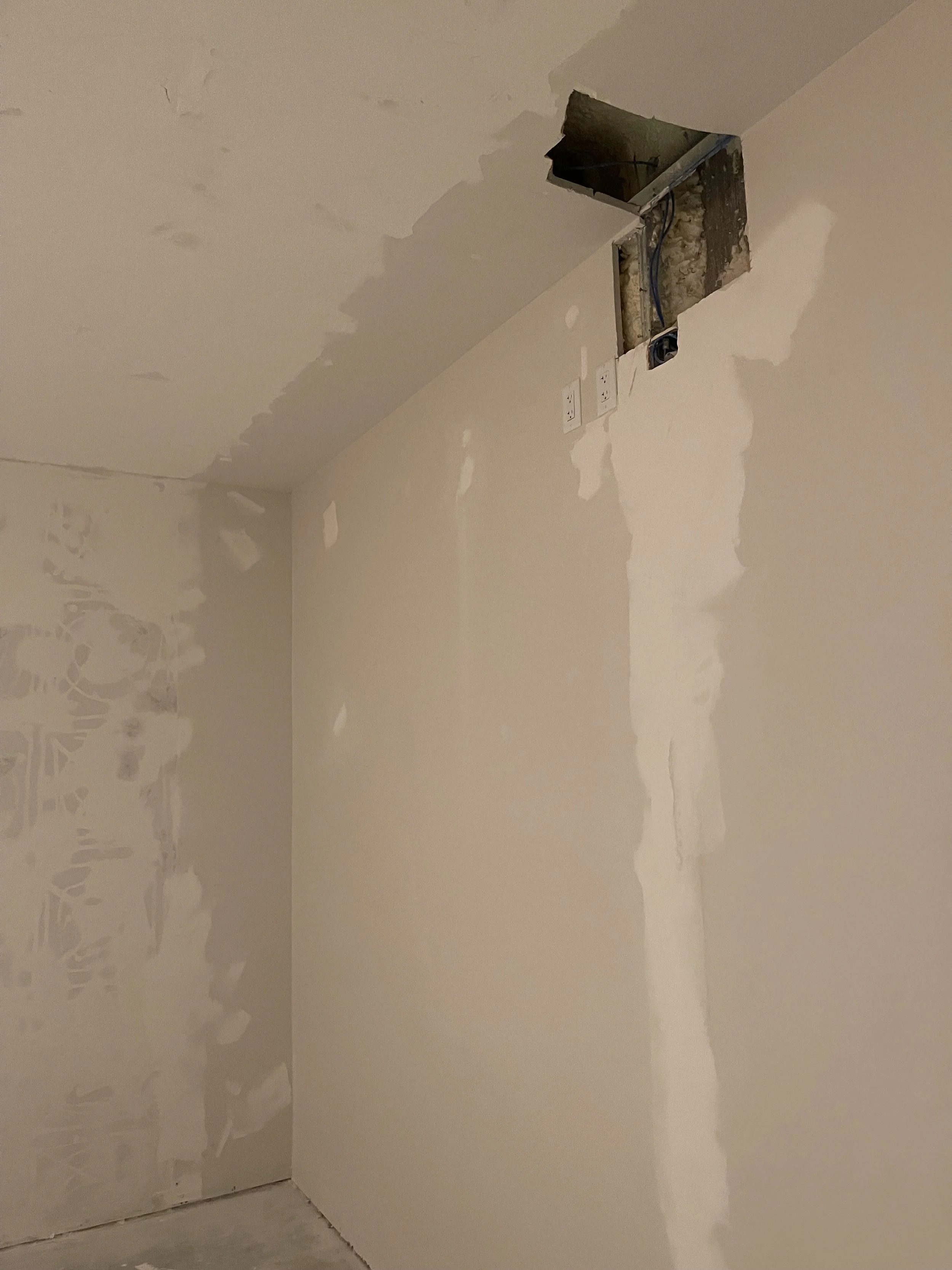 Unfinished wall with drywall on a corner, some patches of mud, and an open rectangular cutout at the ceiling showing electrical wiring and an electrical outlet.