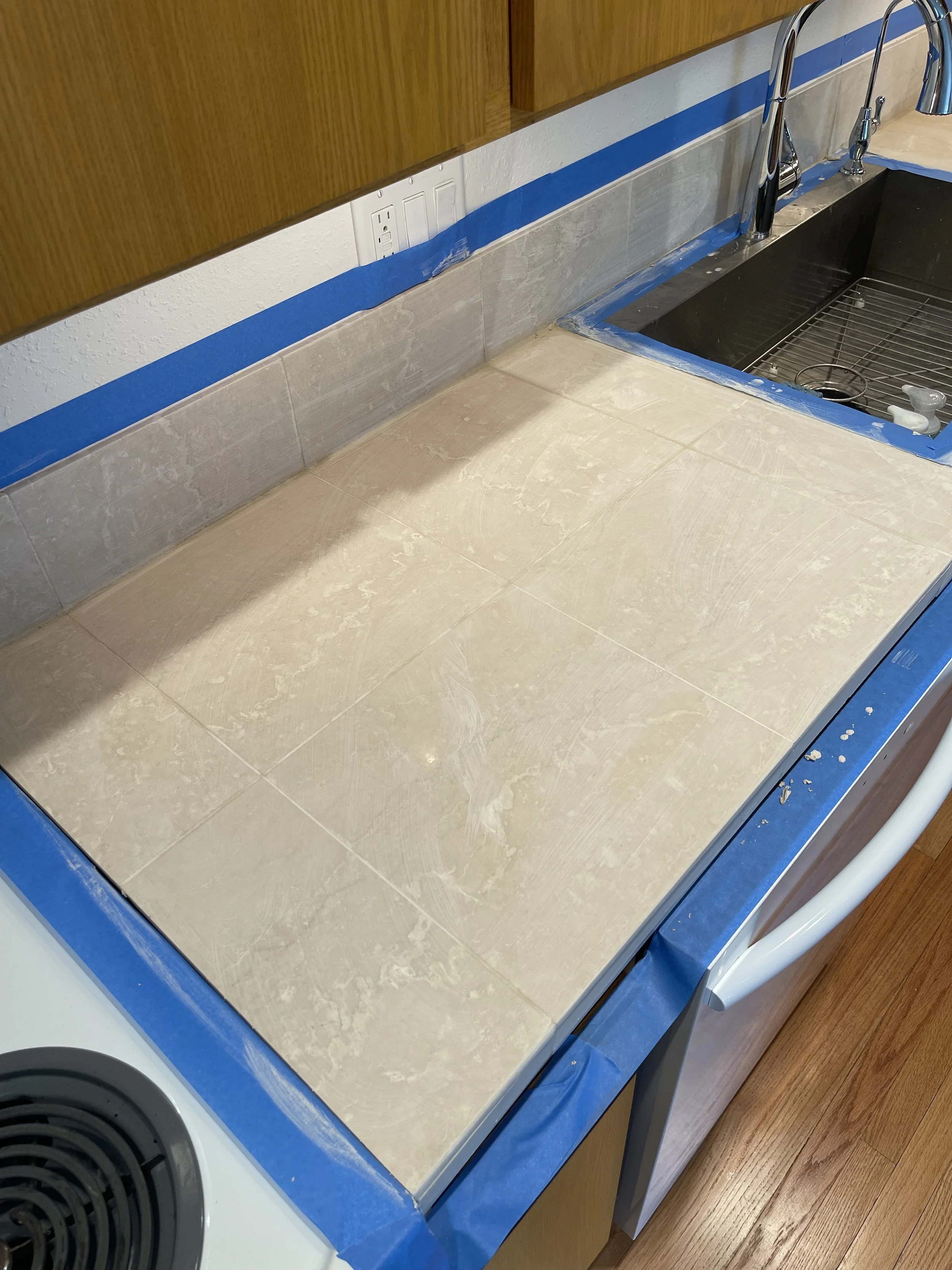 Kitchen countertop under renovation with beige tiles partially installed, blue painter's tape along edges, and a black sink with a chrome faucet.