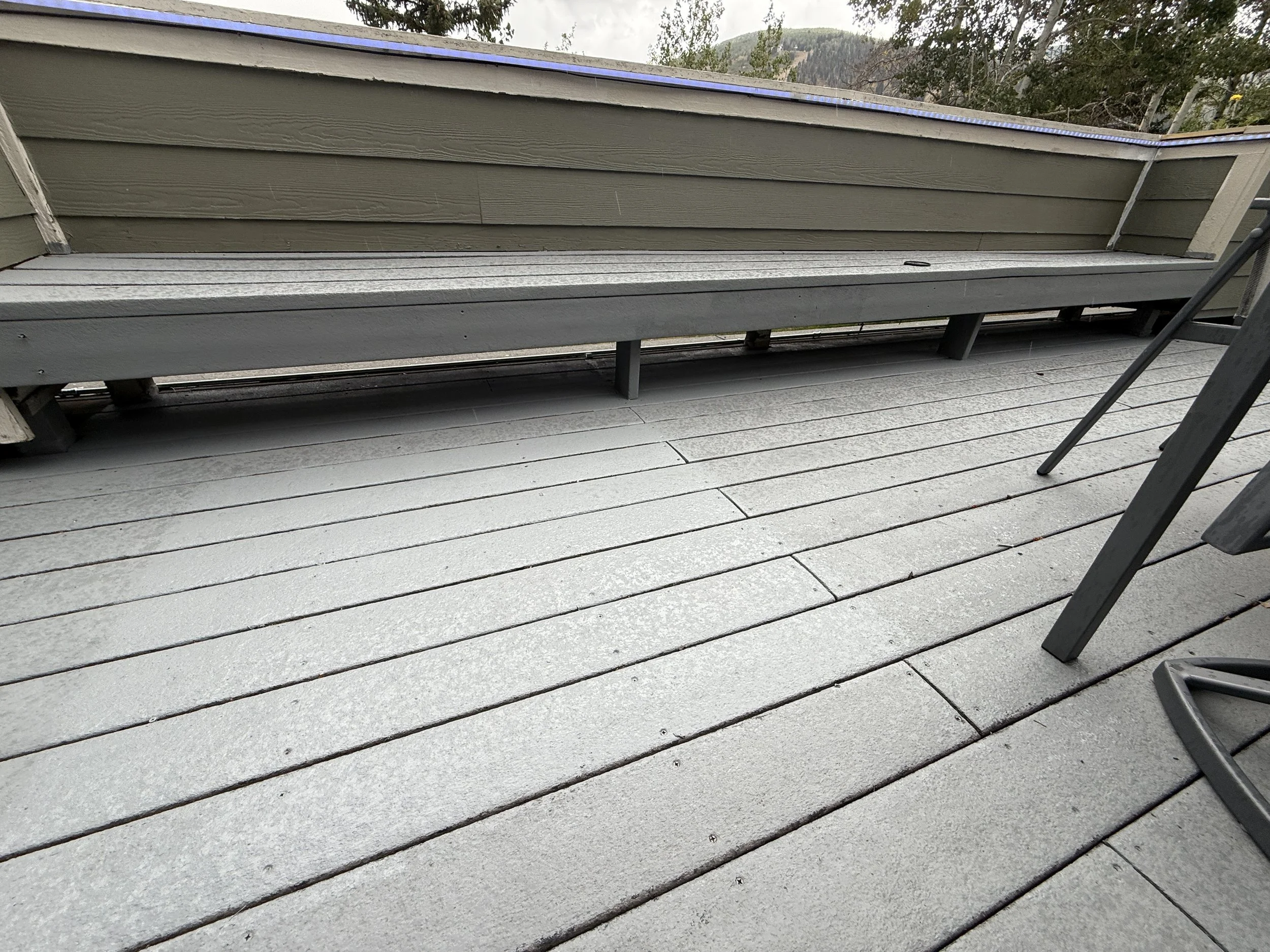 A gray wooden deck with a bench along the house wall and a metal chair to the right.