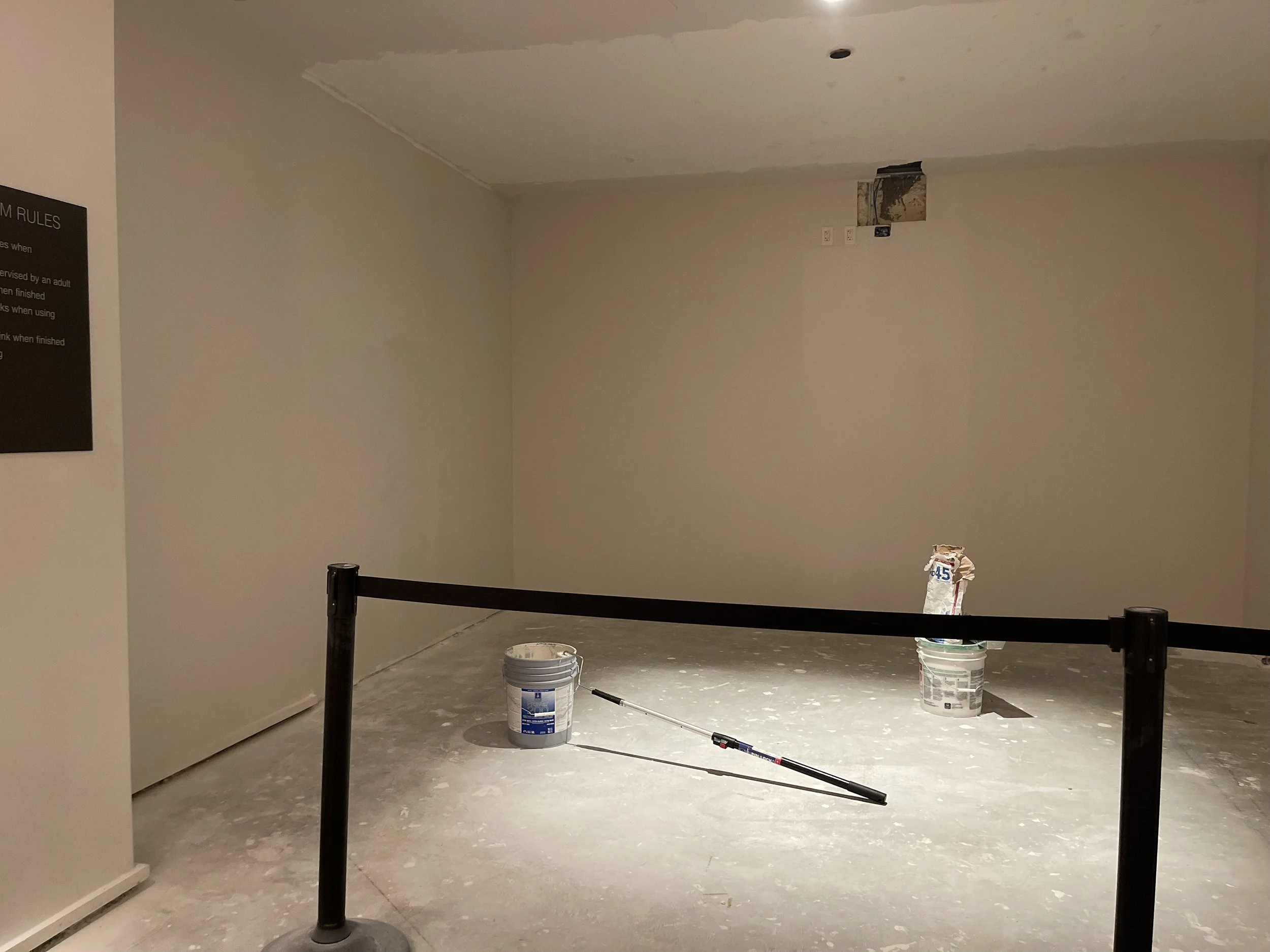 Construction area with unfinished wall and ceiling, a bucket, a bag of drywall compound, and a paint roller on the floor, surrounded by black ropes.