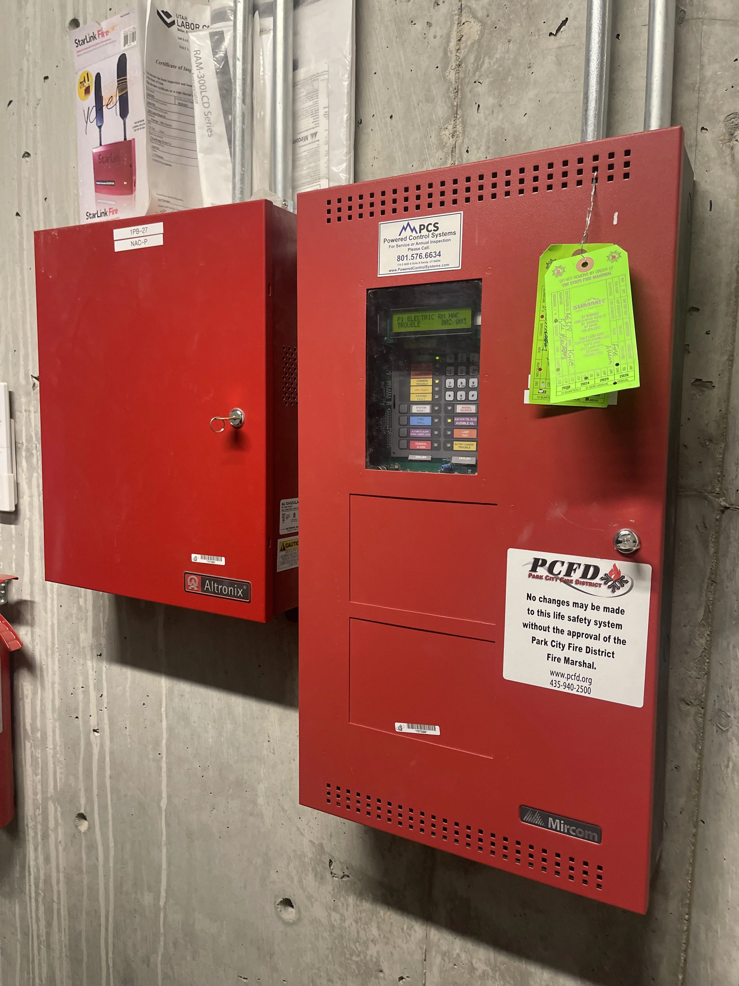Red fire alarm control panel and red fire alarm cabinet mounted on a concrete wall with informational labels and tags.