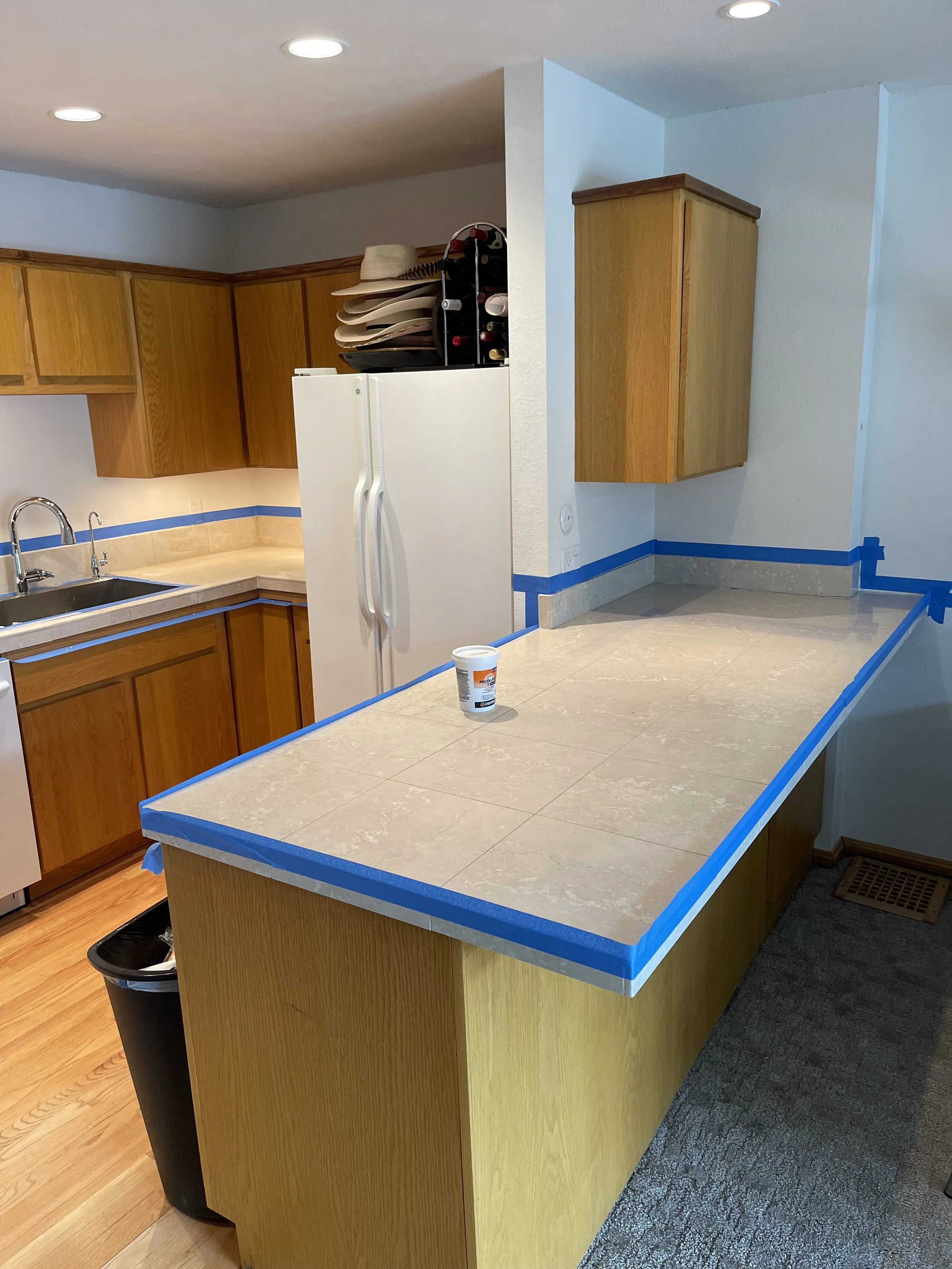 A kitchen with wooden cabinets, beige countertop, white refrigerator, and a tiled breakfast bar with painter's tape around the edges. There is a container on the counter, a trash can underneath, and some hats and wine bottles on top of the refrigerat