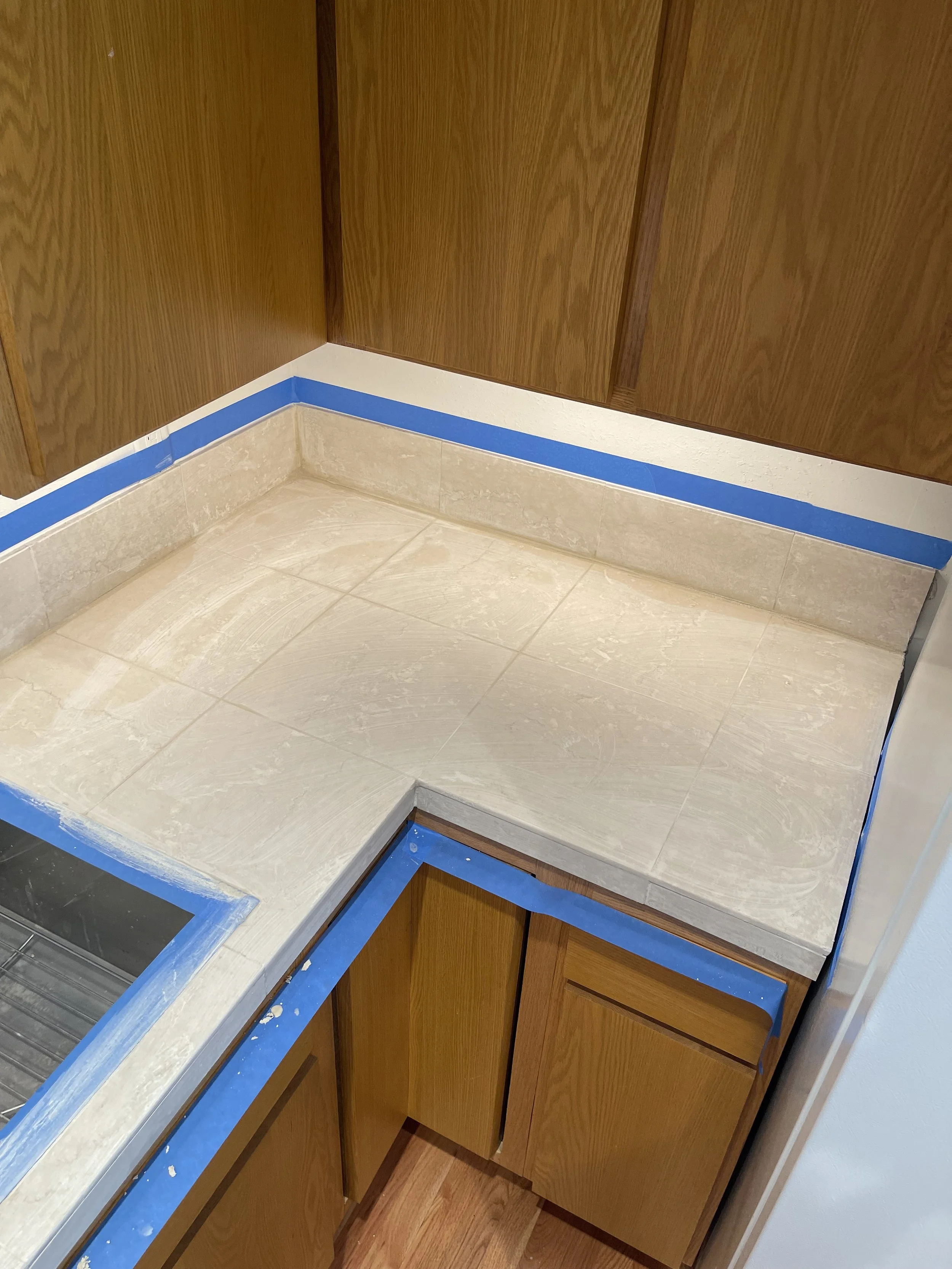 Kitchen corner with new beige tile countertop and wooden cabinets, blue painter's tape along the edges to protect surfaces during renovation.