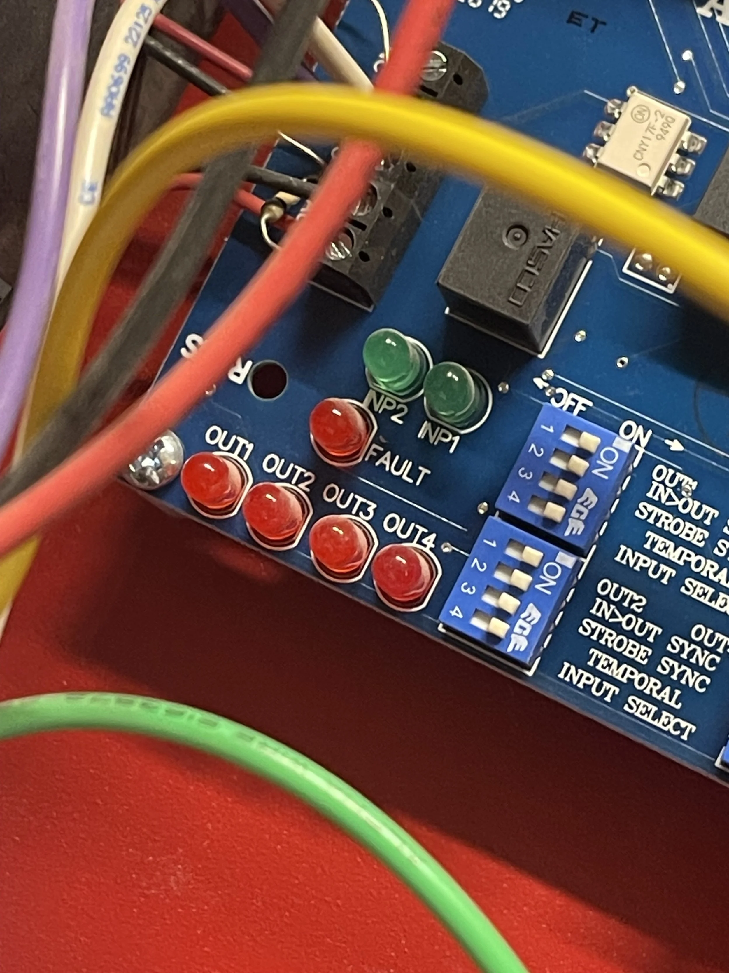 Close-up of a blue electronic circuit board with several red, green, and purple wires connected. There are various LEDs, switches, and components such as relays and pin headers, with some labeled outputs and functions.