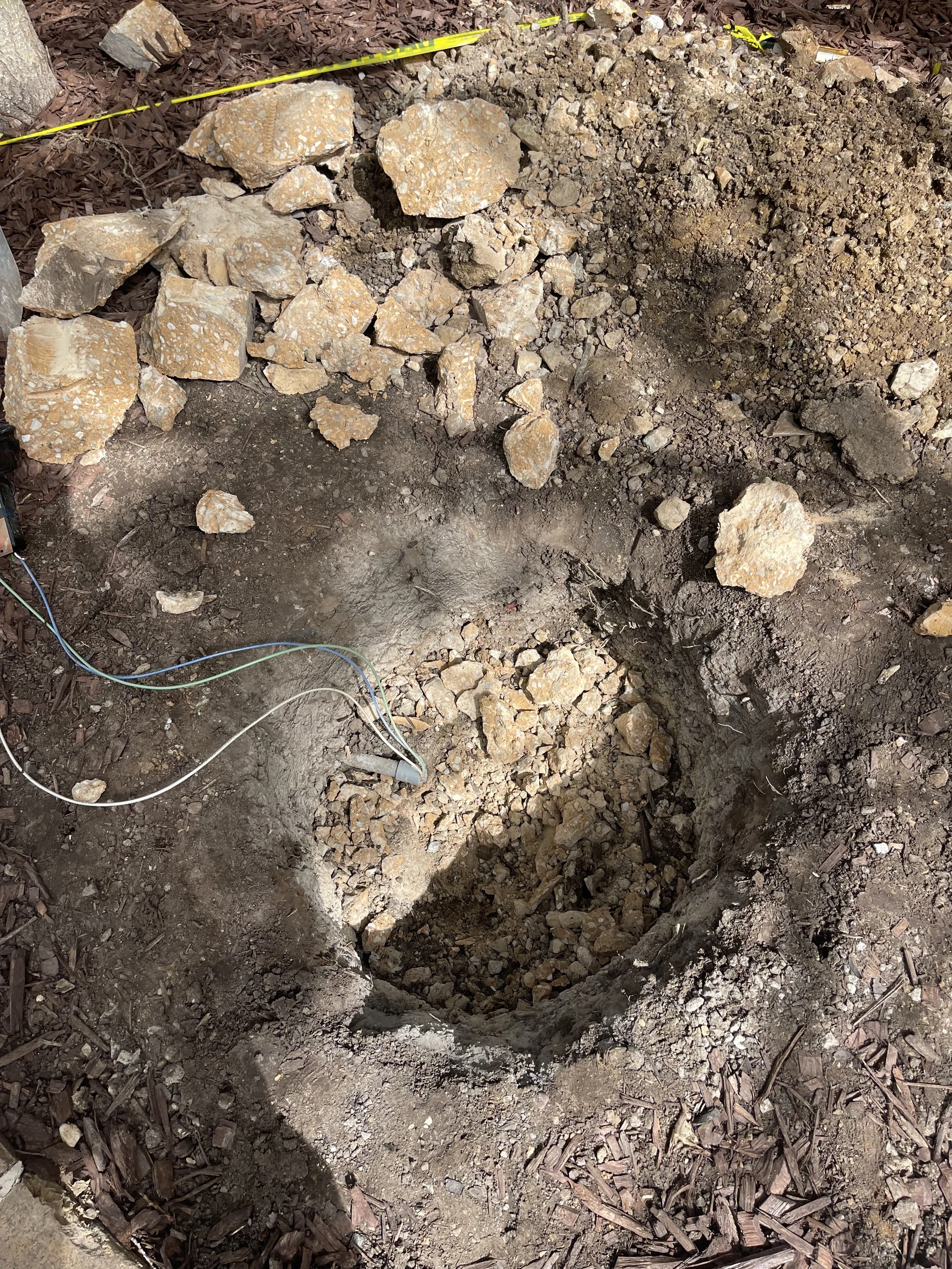 A hole in the ground surrounded by broken rocks and dirt, with exposed wires near the edge, likely part of an excavation or construction site.