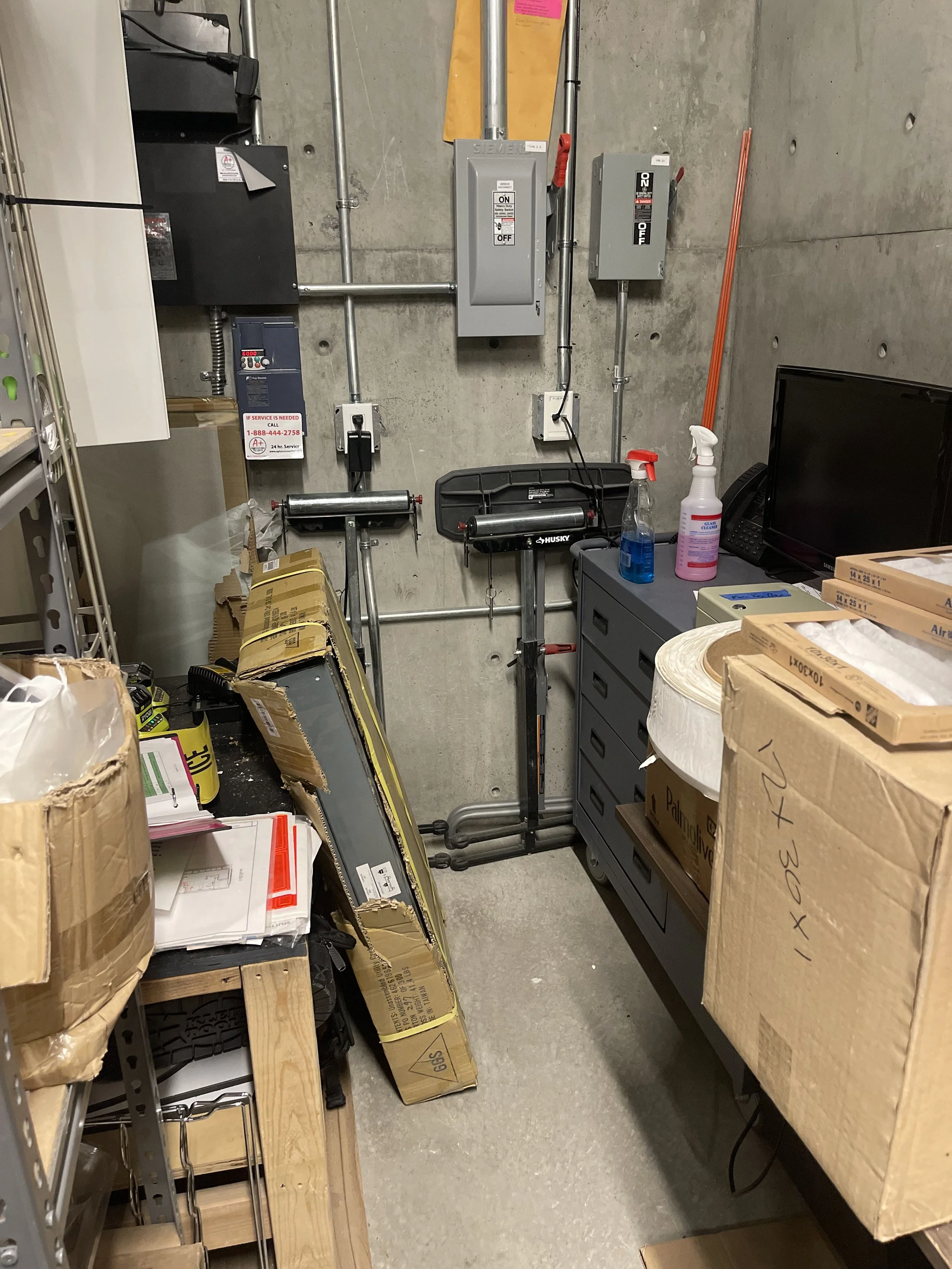 A cluttered storage room with boxes, a workbench, cleaning supplies, and electrical panels on a concrete wall.