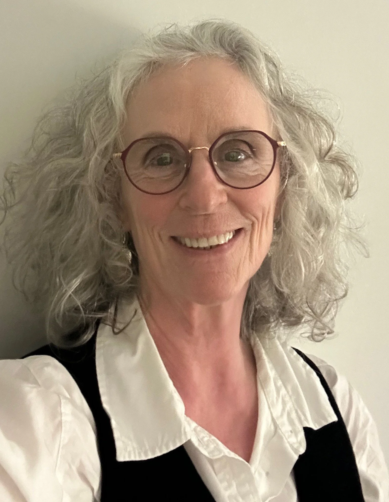 A smiling woman with curly gray hair and glasses wearing a white blouse and black vest.
