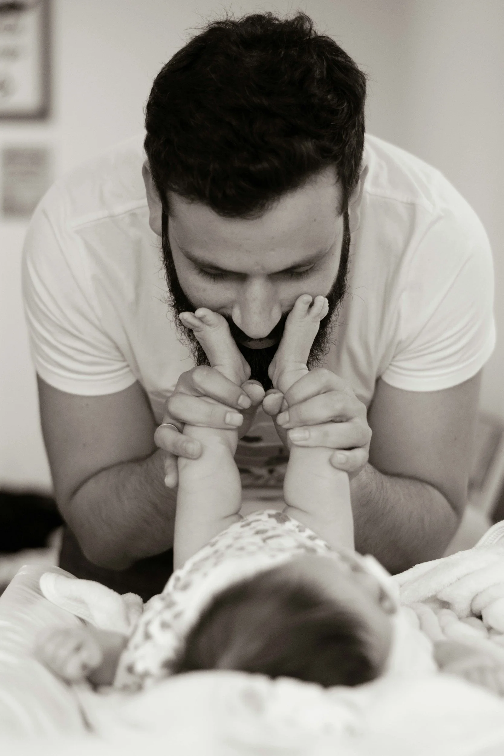 A man with a beard holds a baby's feet, leaning over the baby who lies on a bed, in a caring and tender moment.