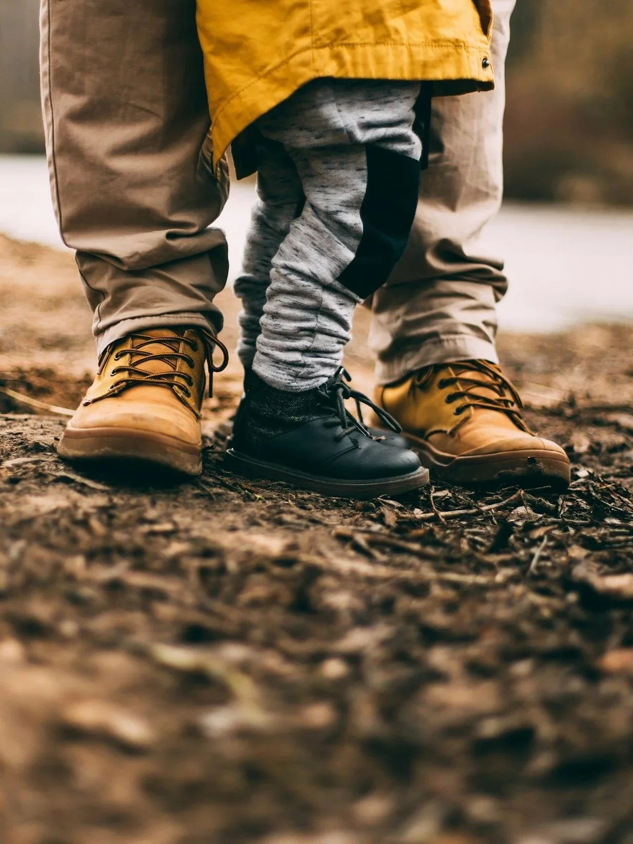 Close-up of a child's legs and feet, with the child wearing one yellow boot and one black boot, standing on a forest trail with brown leaves.