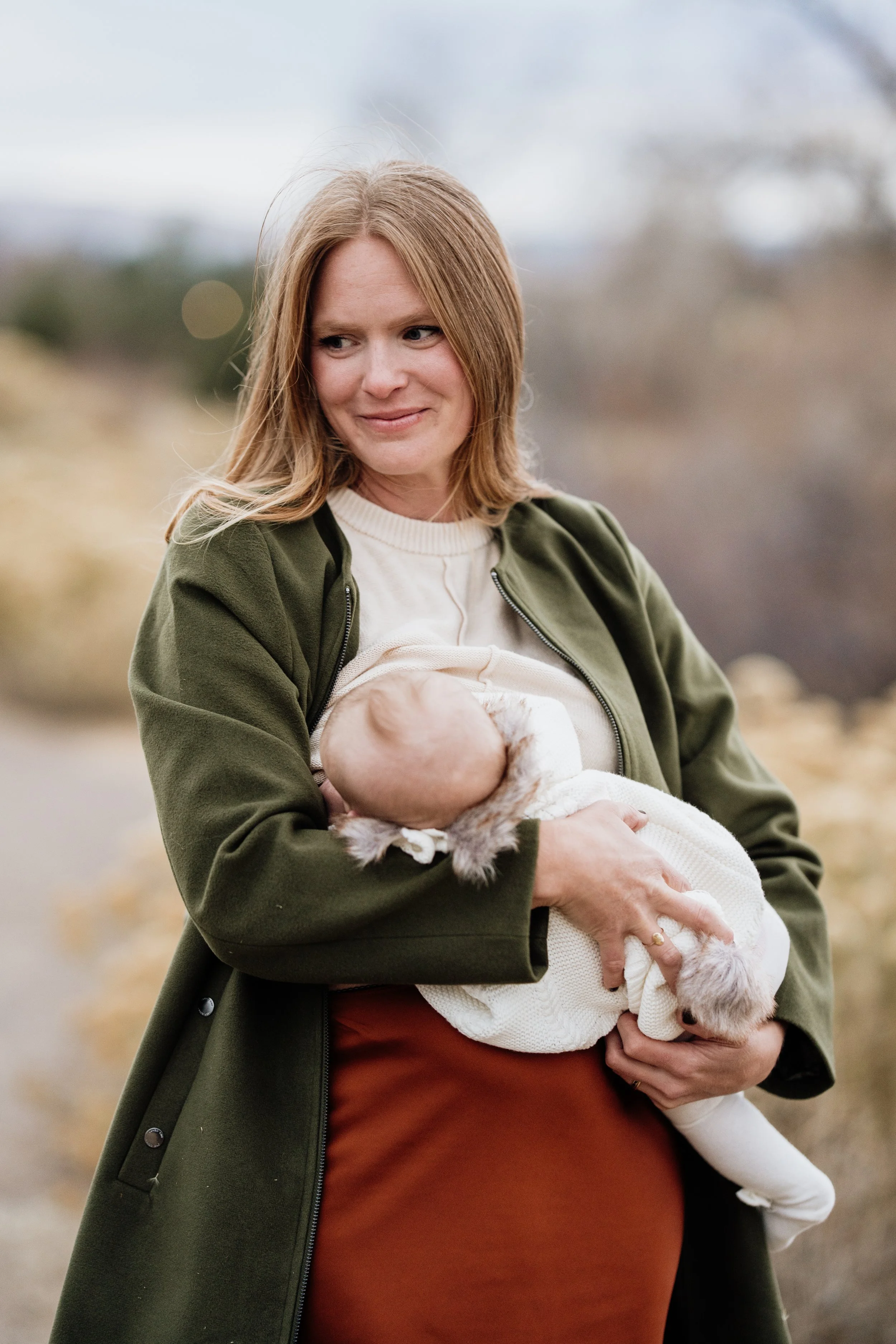 A woman with shoulder-length red hair, wearing a green coat and a cream-colored sweater, is holding a baby wrapped in a white blanket outdoors.
