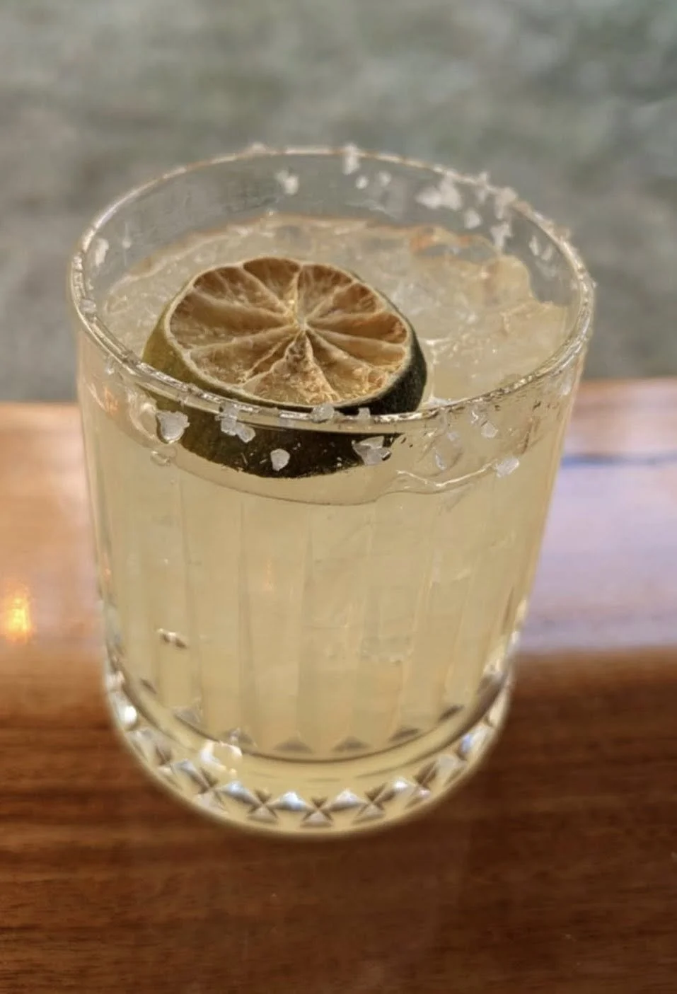 A clear glass with a salted rim, filled with a yellowish beverage, garnished with a slice of dried lemon or lime.