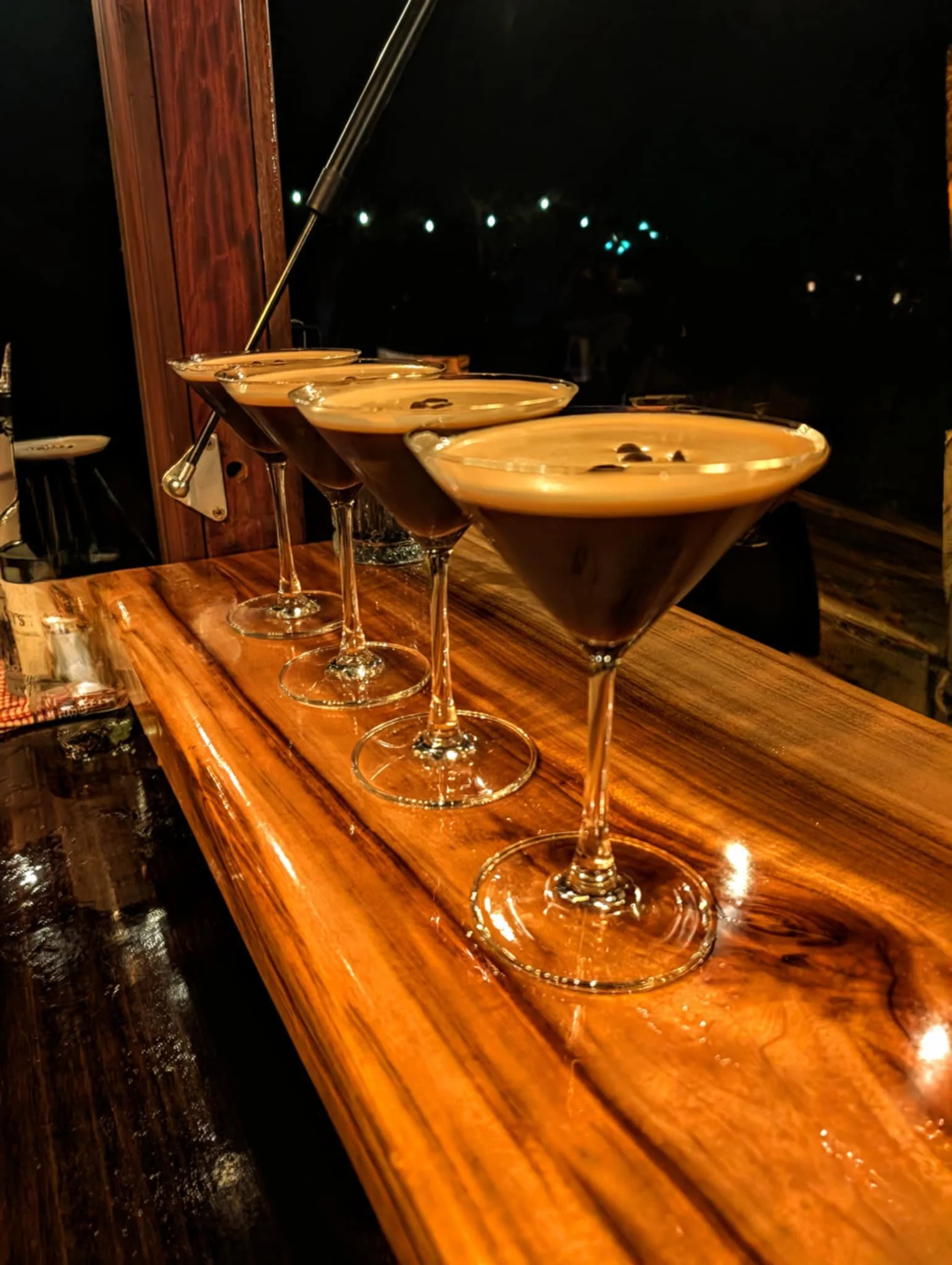Four cocktails in martini glasses lined up on a wooden bar counter at night.