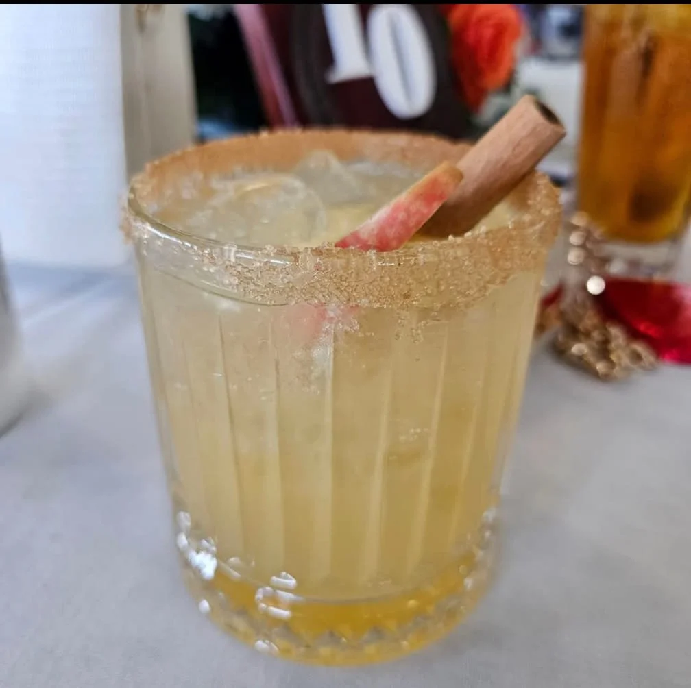 A glass of a yellow beverage with ice and a cinnamon stick, rimmed with sugar.