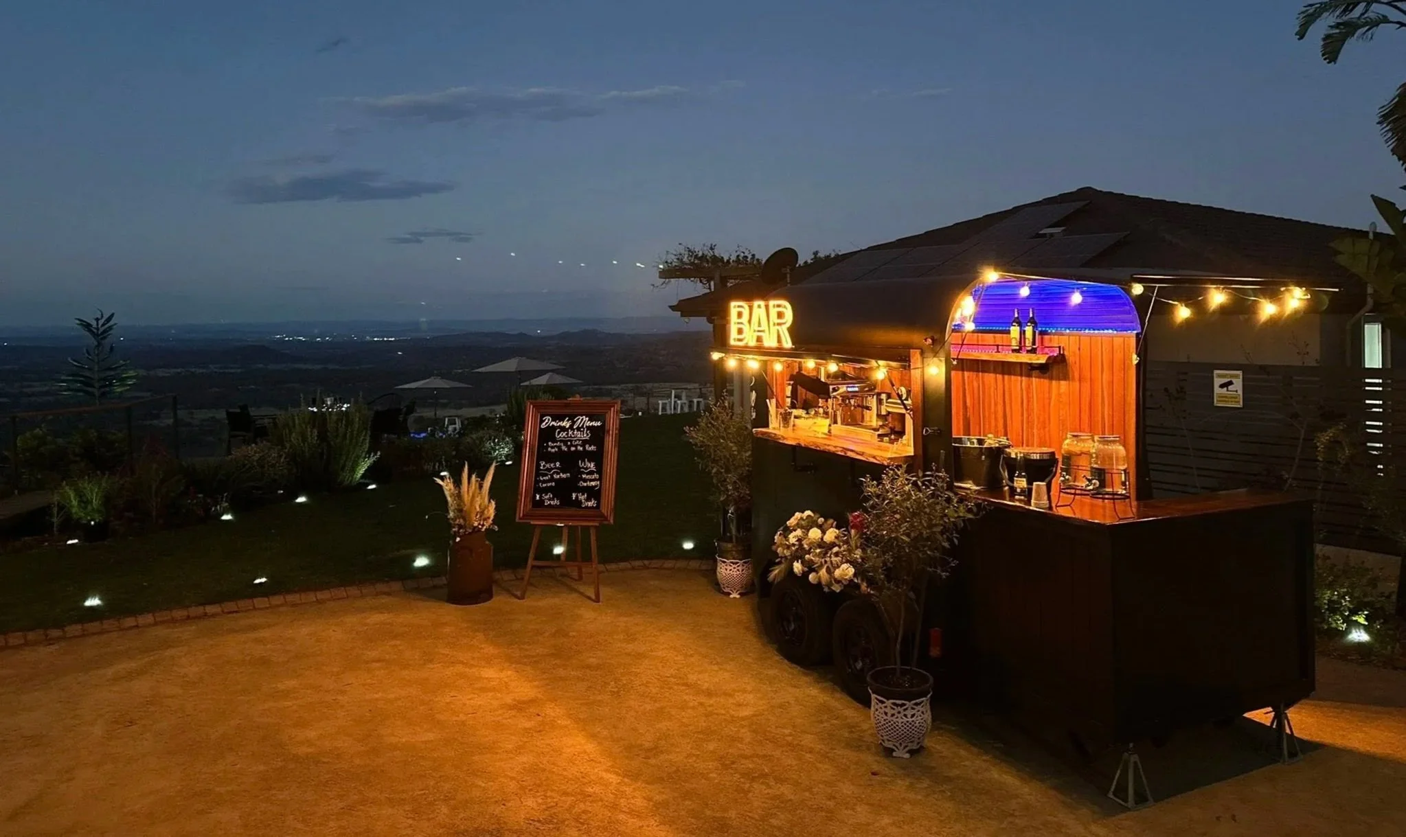 A mobile bar set up outdoors on a grass area at dusk, with a mountain landscape in the background, decorated with string lights and flowers.