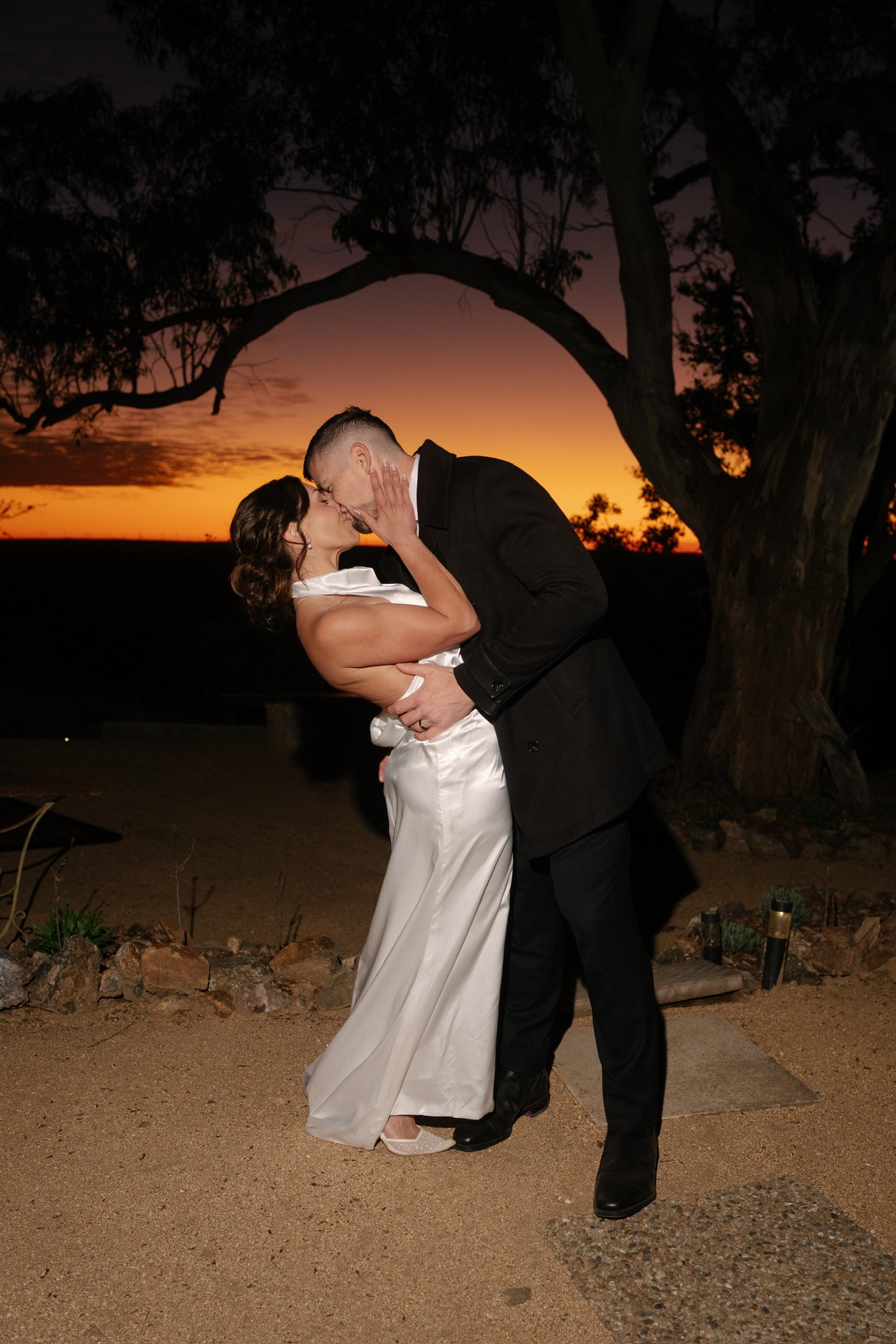 A newlywed couple embracing outdoors, smiling happily. The woman wears a lace wedding dress and a flower crown, and the man is in a black suit with a bow tie.