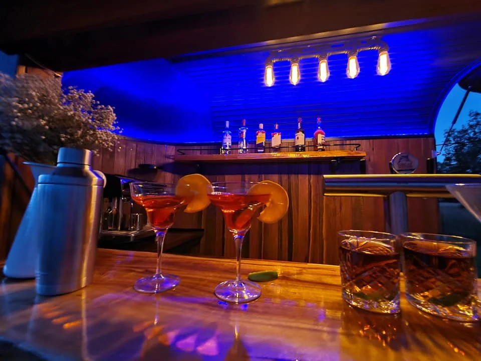 A bar scene with two cocktails garnished with orange slices on a wooden table, two glasses of whiskey, and various bar supplies under warm lighting with a blue-lit ceiling.