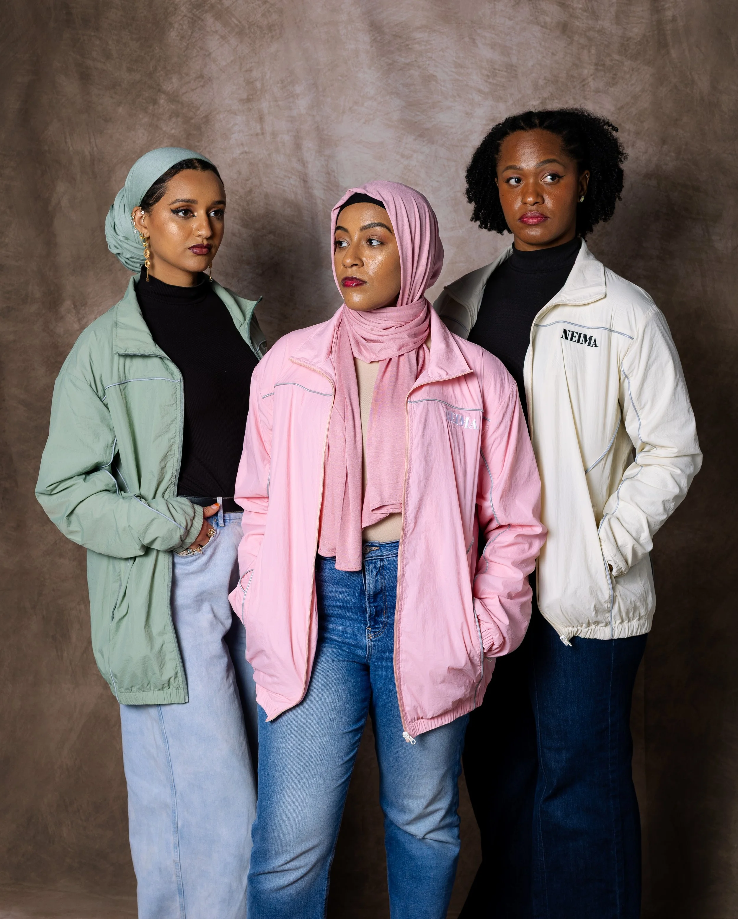 Three women standing against a neutral background, wearing casual jackets and jeans, with two wearing hijabs and one with natural curly hair.