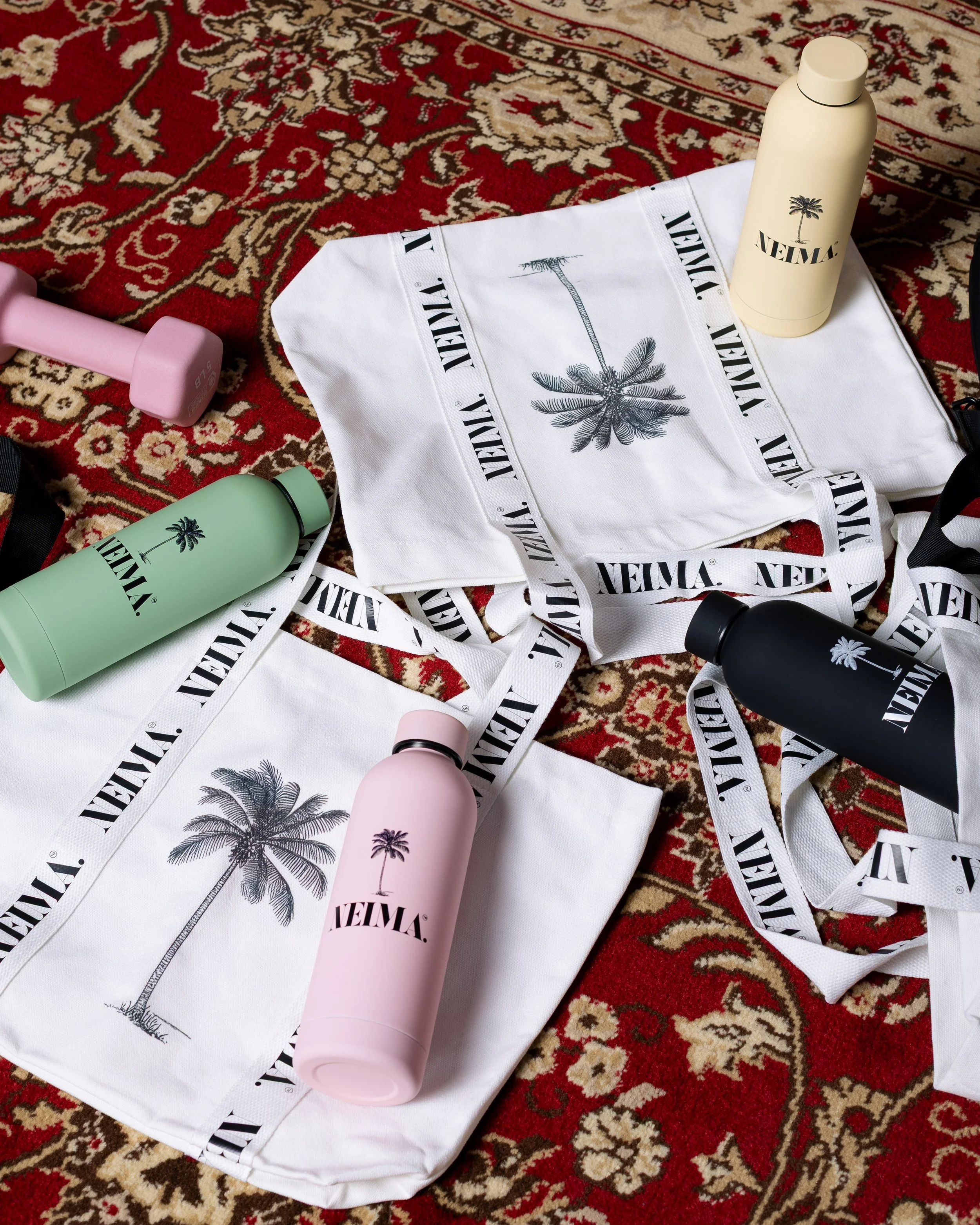 Colorful reusable water bottles, tote bags with palm tree designs, and a pink dumbbell on a red patterned carpet.