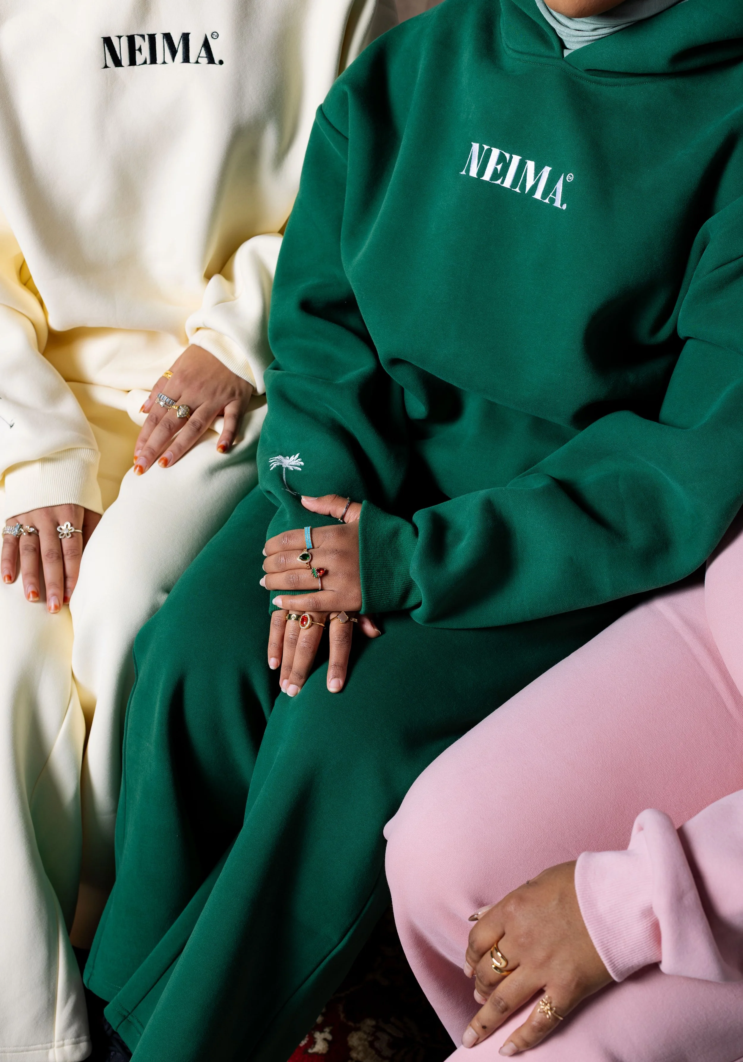 Close-up of three individuals wearing colorful sweatshirts and pants, with hands adorned with rings resting on their laps. The sweatshirts have the word 'NEIMA' printed on them, and one sweatshirt features a small palm tree embroidery.