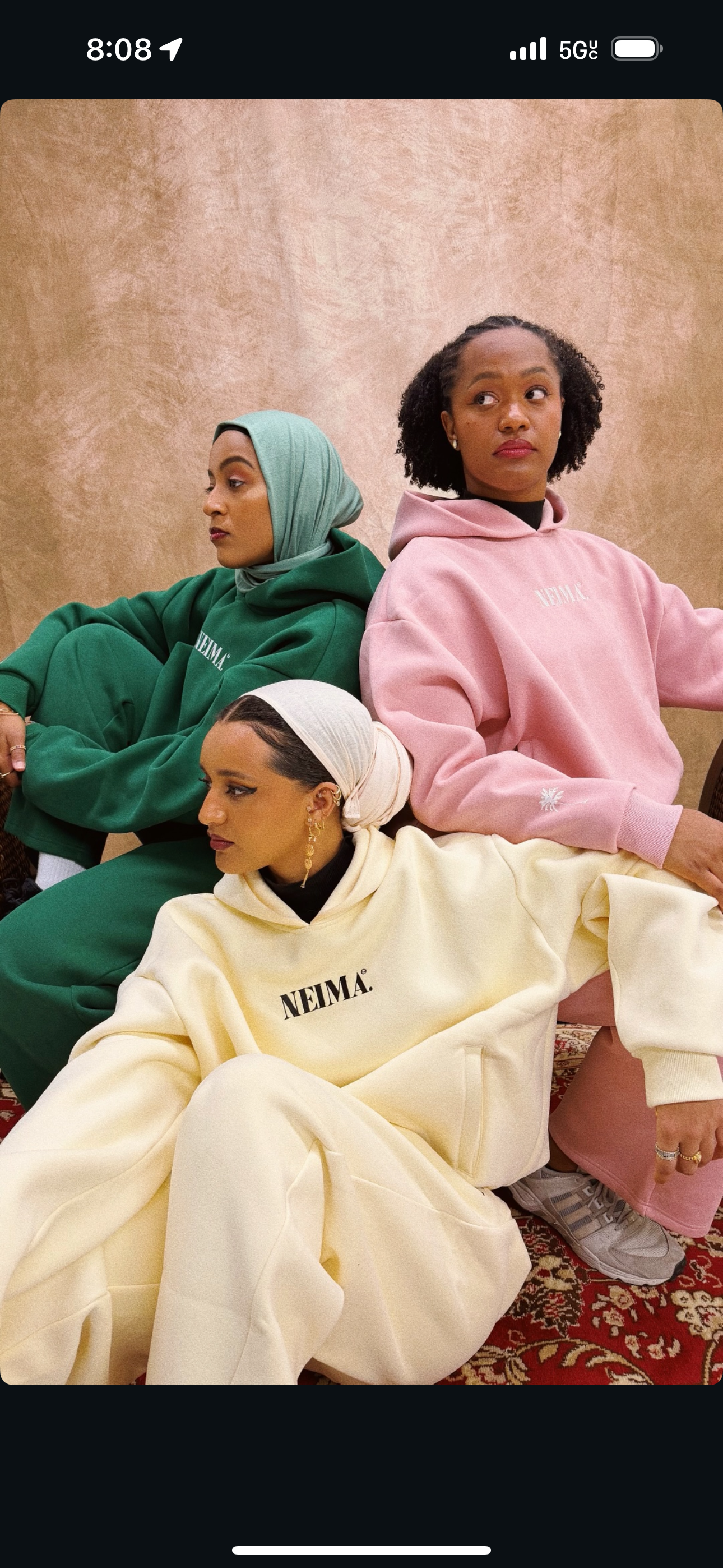 Three women wearing hoodies with the word NEIMA printed on them, sitting against a tan wall and a patterned carpet.