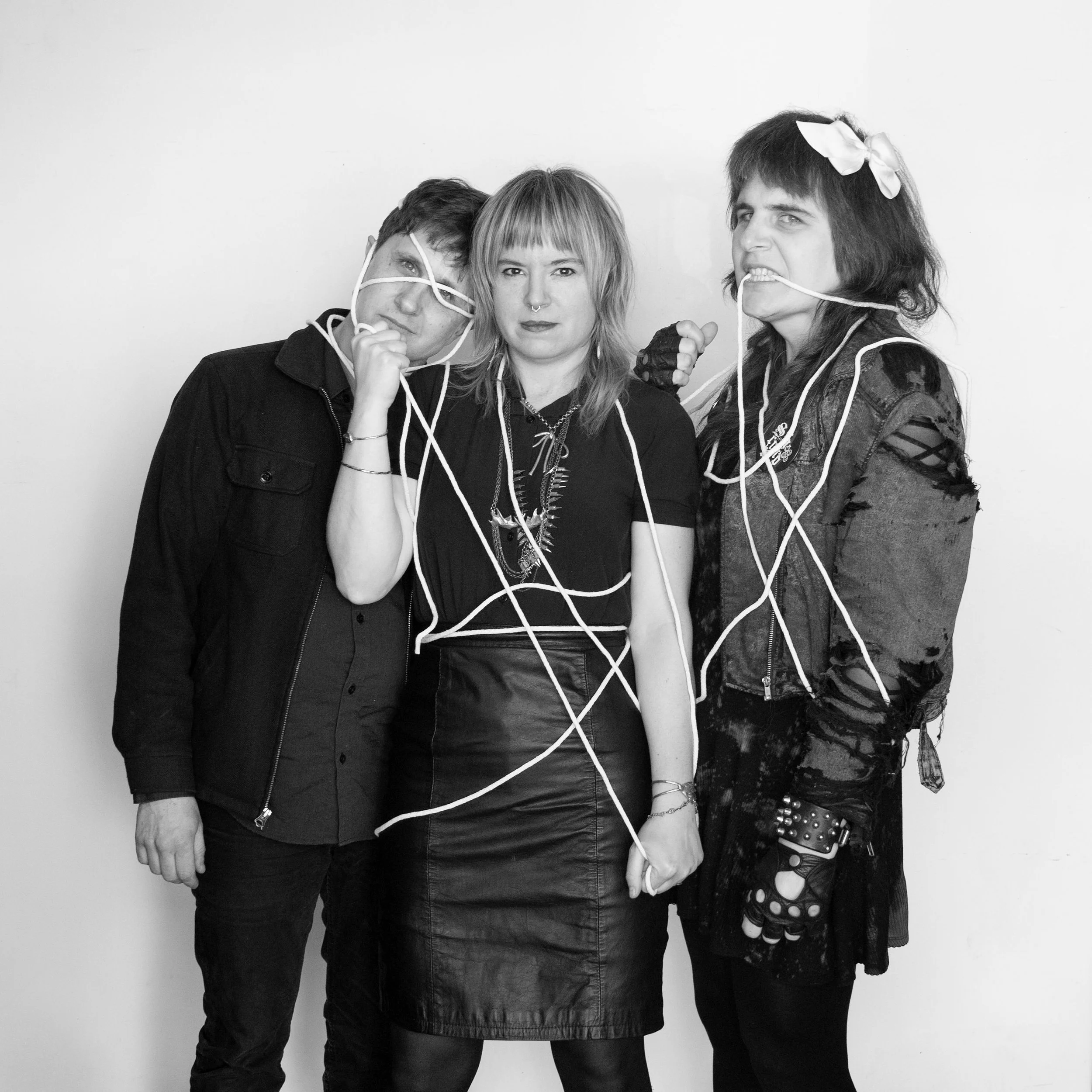 Three people standing together in a black and white photo, tangled in white cords, with two women and one man, all with punk/alternative fashion and hairstyles, posing against a plain background.