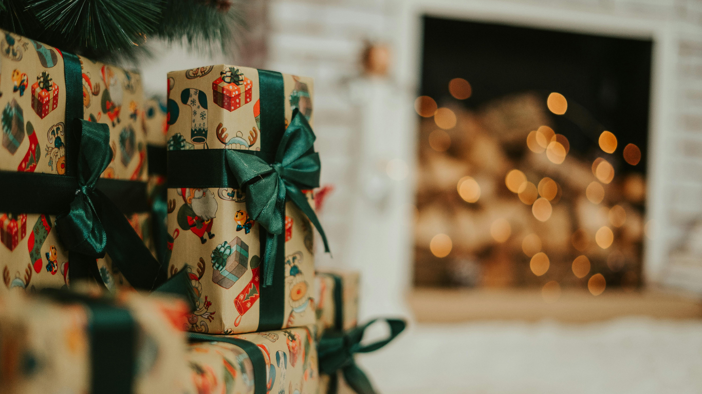 Close-up of Christmas presents wrapped in festive paper with green ribbons under a decorated Christmas tree, with a fireplace in the background.