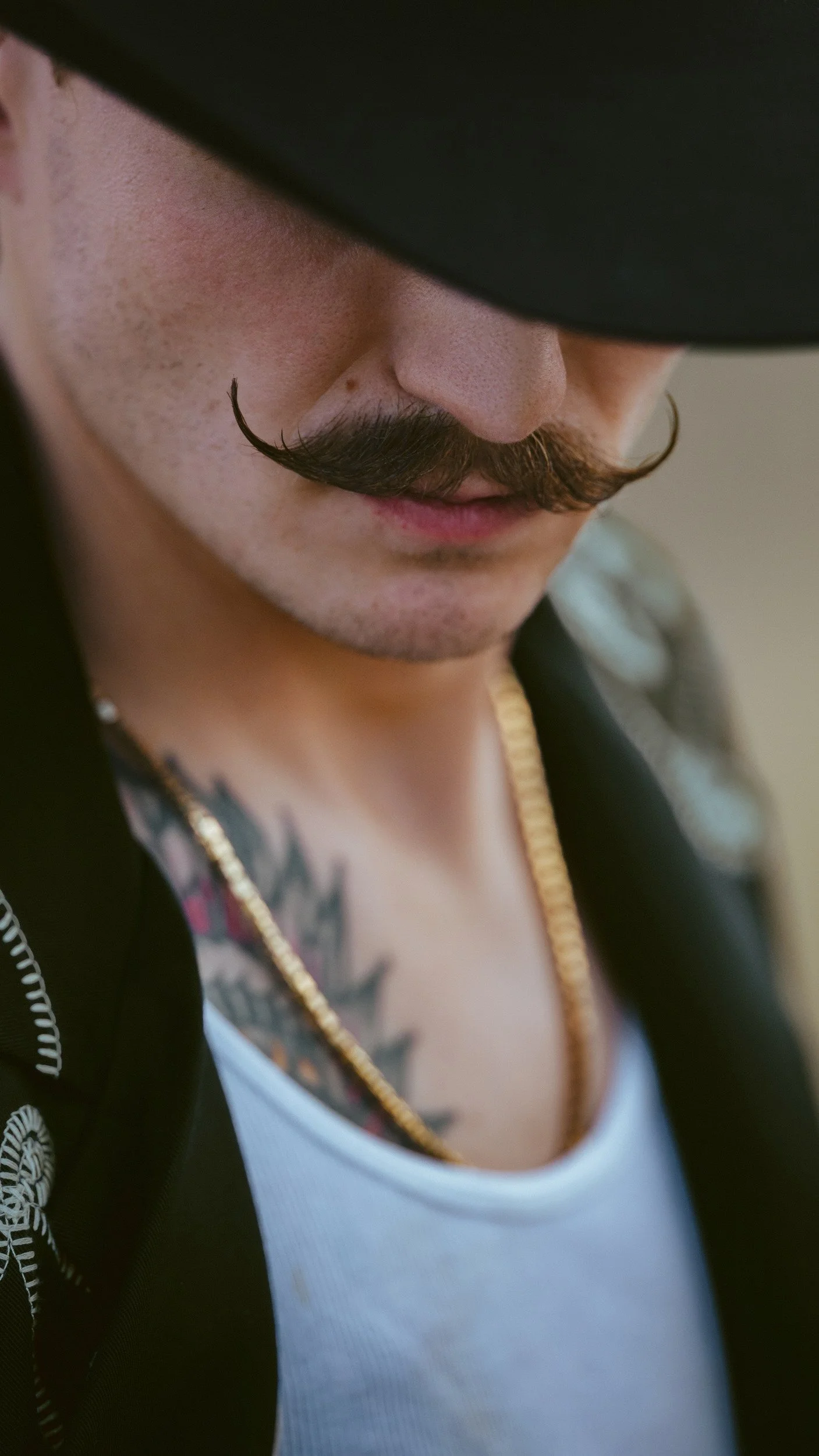Close under the brim of a black hat, a curled moustache, a gold chain and a tattoo above a white tank top and black jacket.
