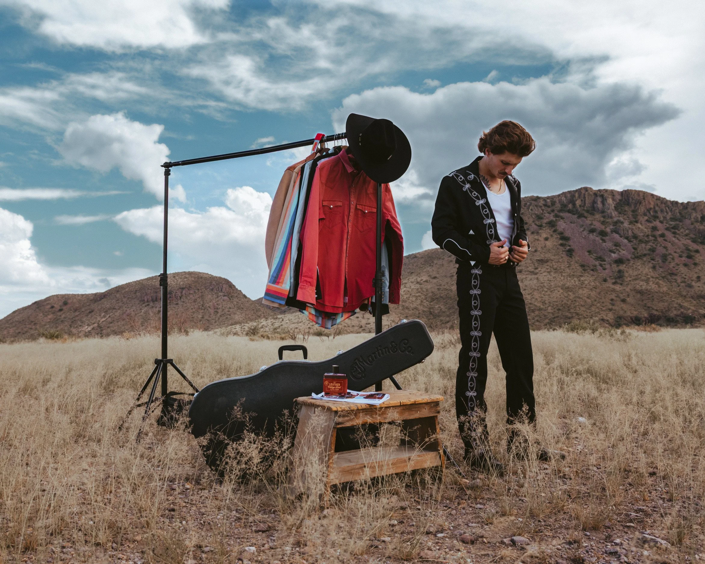 On set in the desert, Braxton Keith buttons a black embroidered jacket beside a rack of western shirts and his guitar case.