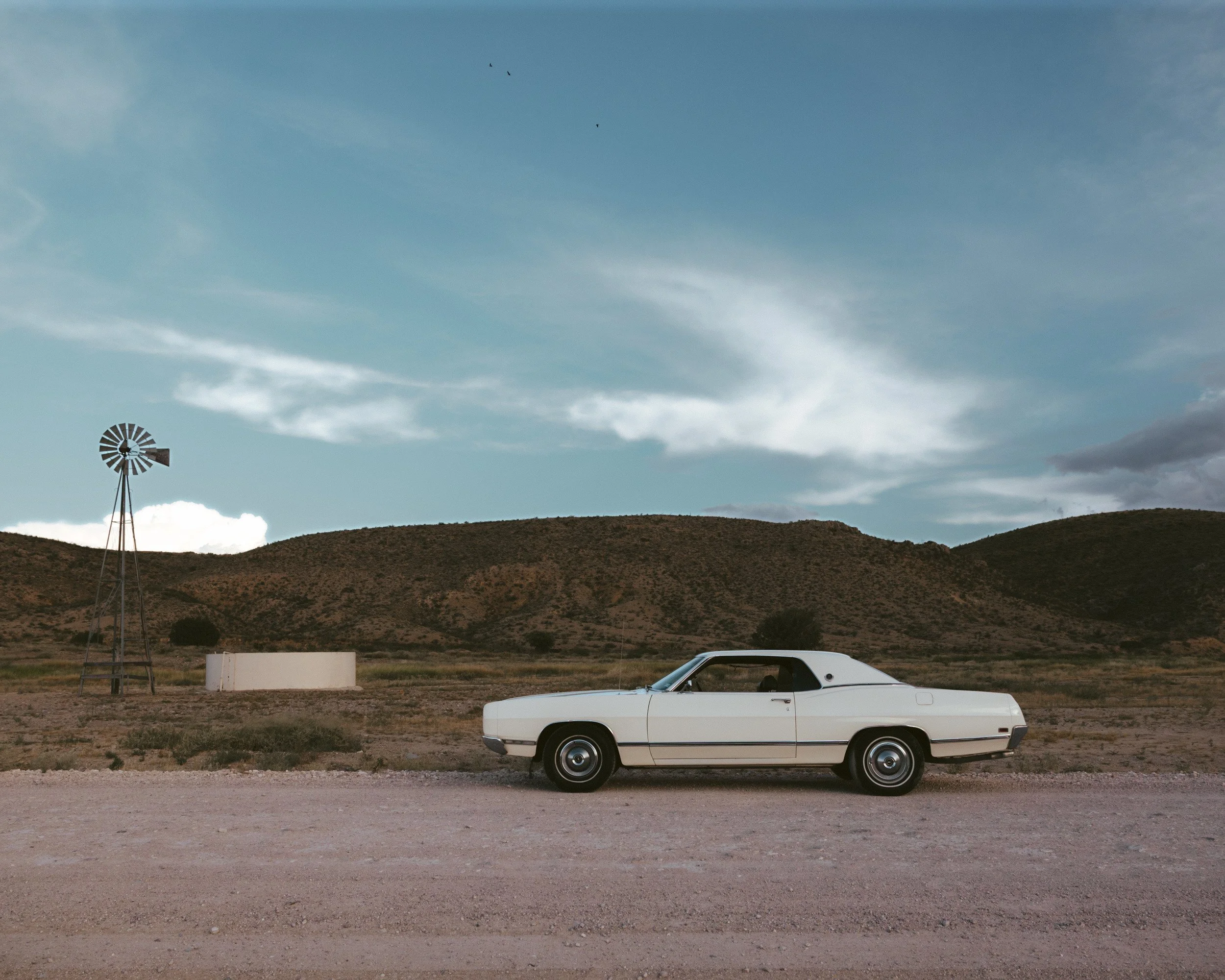 A white vintage car parked on a gravel road beside a windmill and water tank, brown hills and a wide blue sky behind.